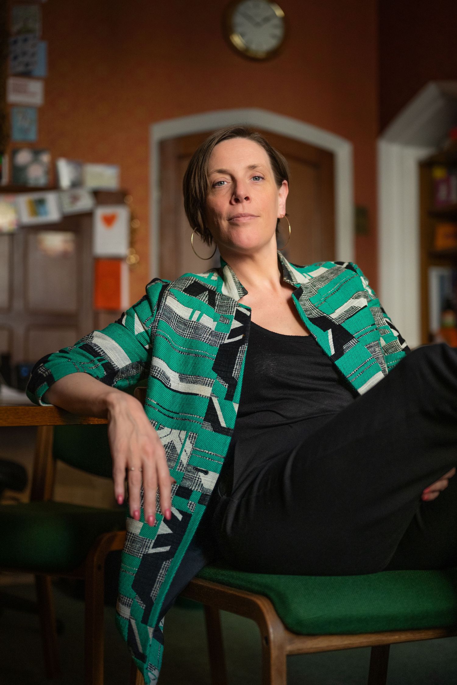 Woman in green geometric patterned blazer sitting relaxed on chair in warm vintage-style office room.