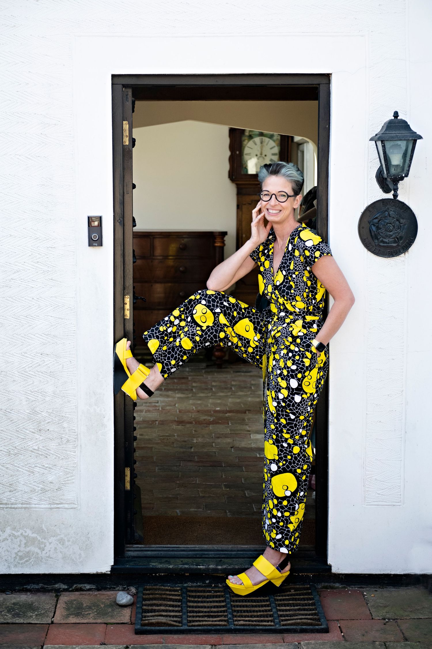 Stylish woman in a black and yellow floral jumpsuit with yellow heels posing in a doorway on the phone.