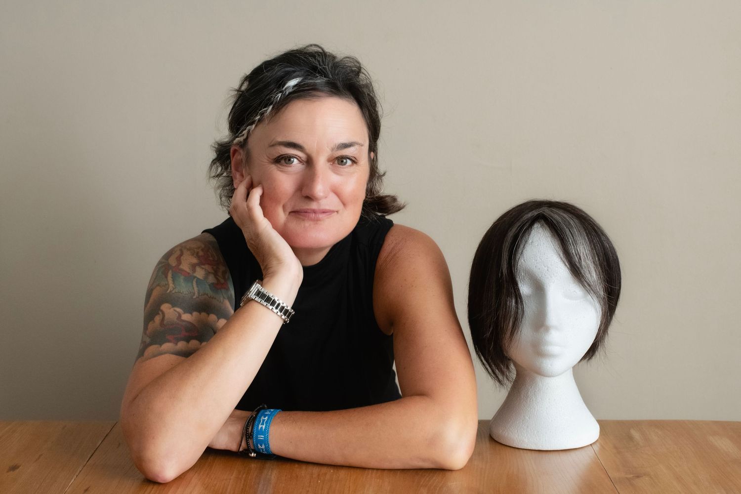 Woman in black top resting chin on hand seated at table with wig on mannequin head.