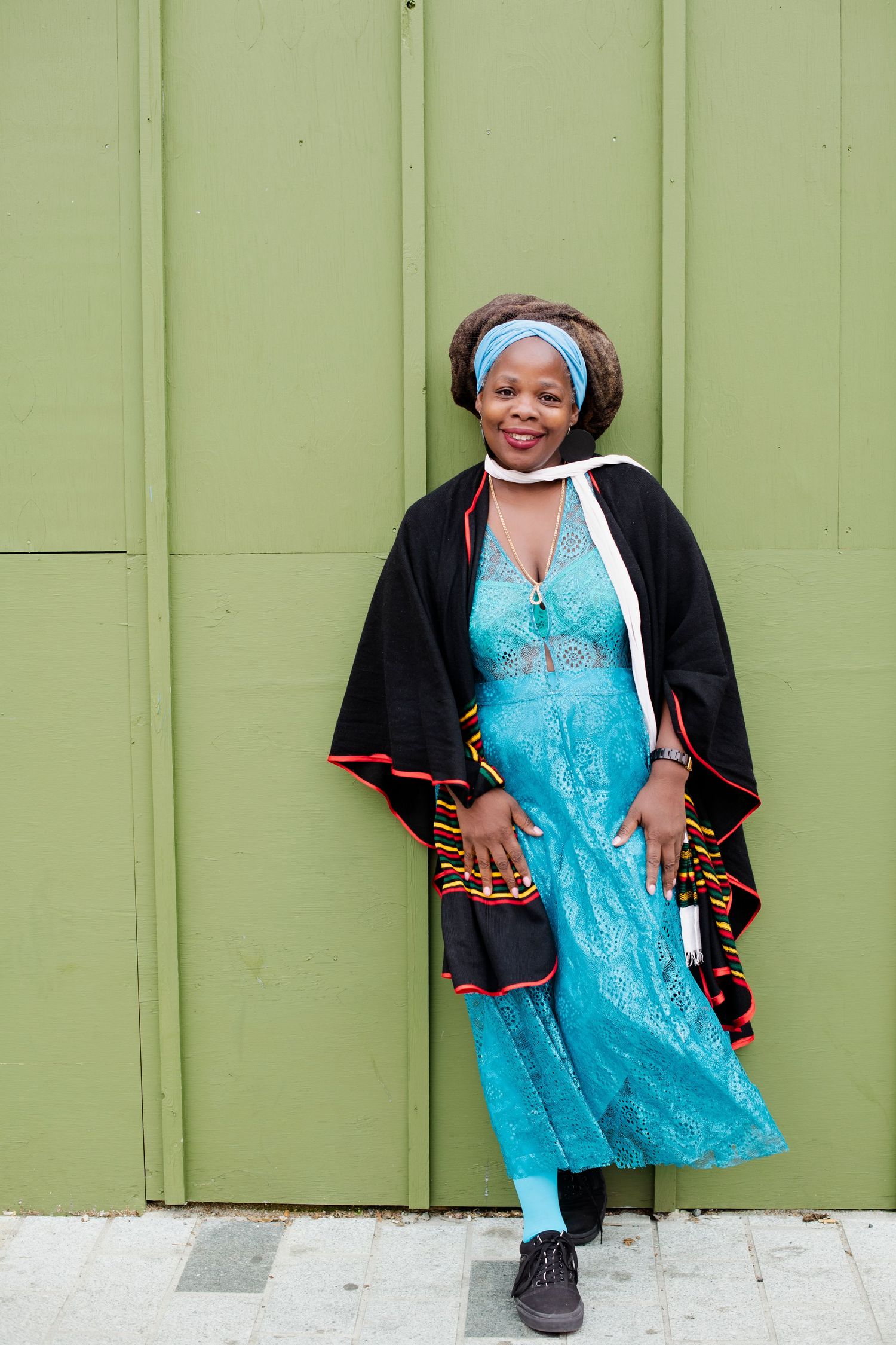 Woman in turquoise lace dress and black African cape with kente trim poses against a green wall.