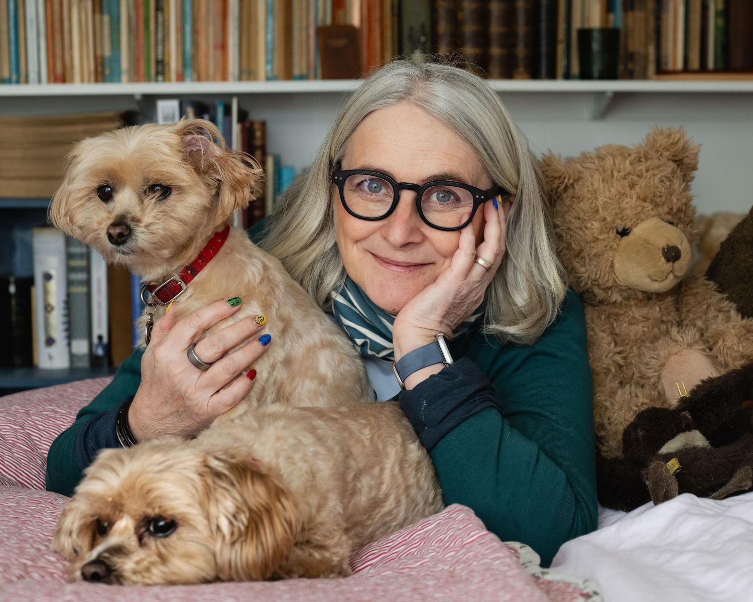 Smiling woman with gray hair and glasses poses with two small dogs and a teddy bear in a cozy library.