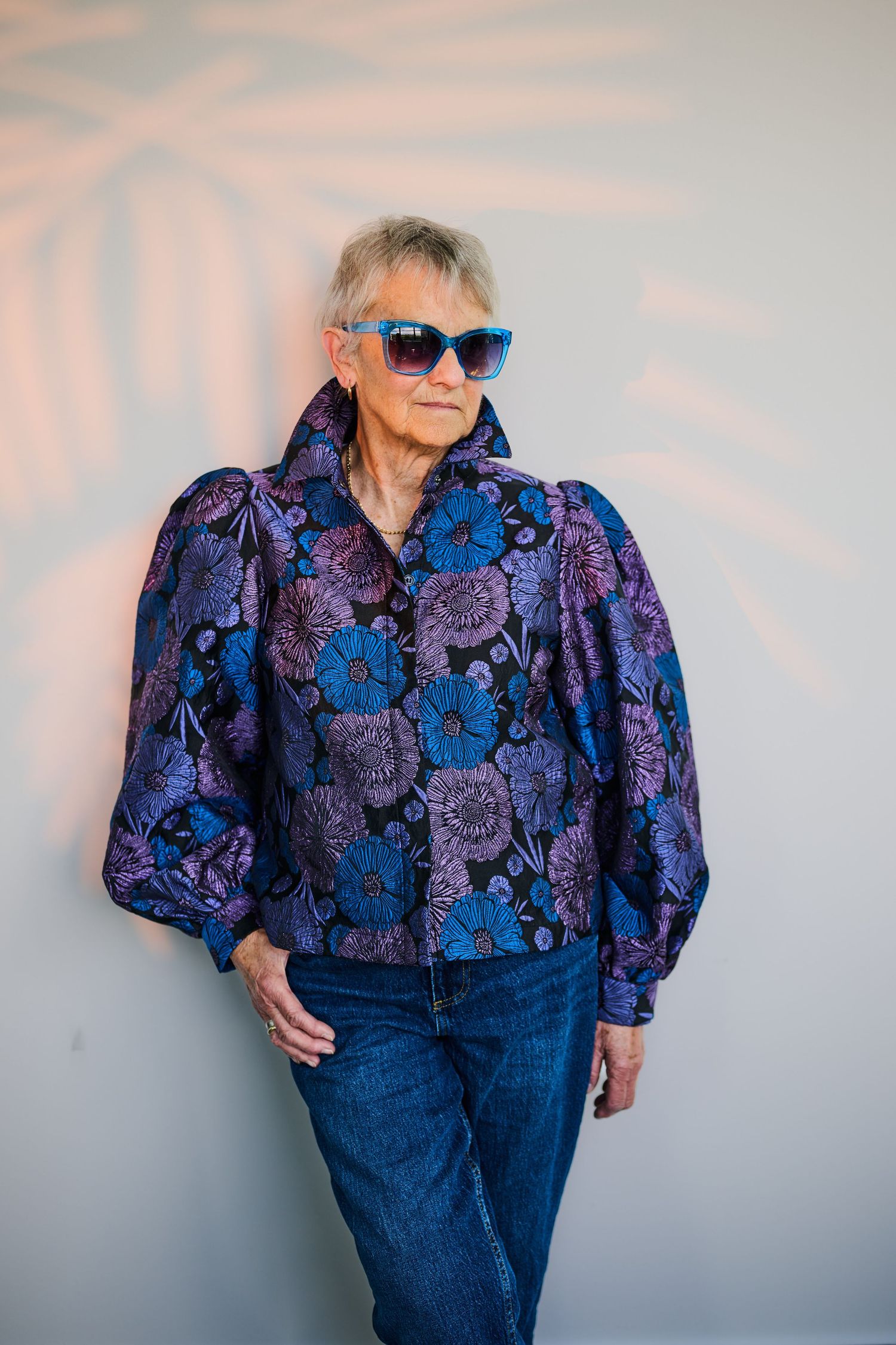 Older woman in blue floral jacket and jeans wearing blue sunglasses, posing confidently against a light gray wall.