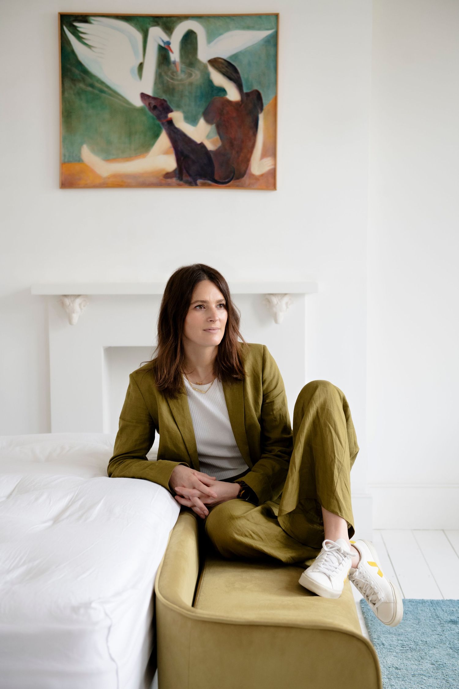 Brunette woman in olive green suit seated on a chair beneath a painting in a white room.