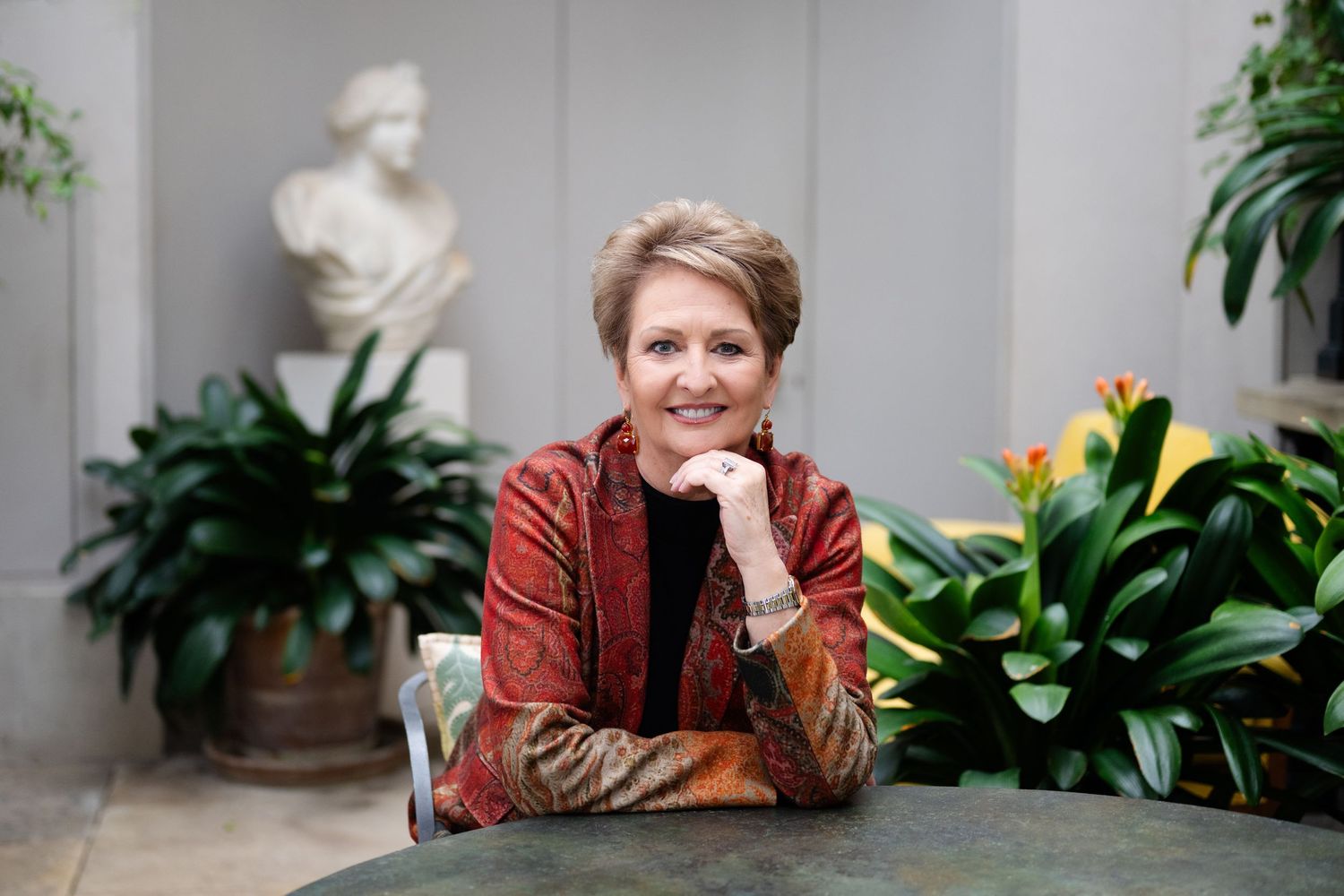 A smiling woman in a red jacket sits at a table surrounded by lush plants and a marble bust sculpture.