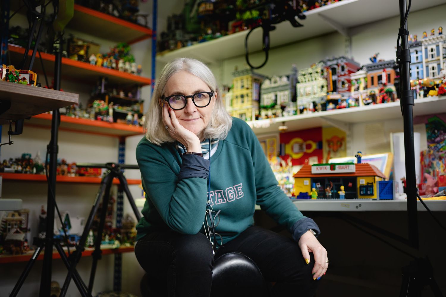 Woman with gray bob and glasses sits thoughtfully among colorful LEGO collection on shelving units.