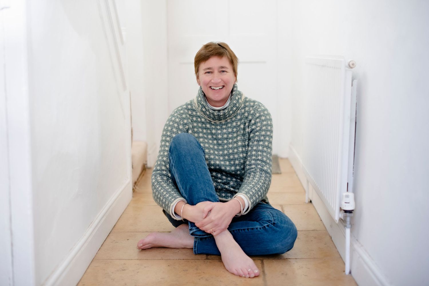 Smiling woman with short auburn hair in a green patterned sweater and jeans sitting on a wooden floor.