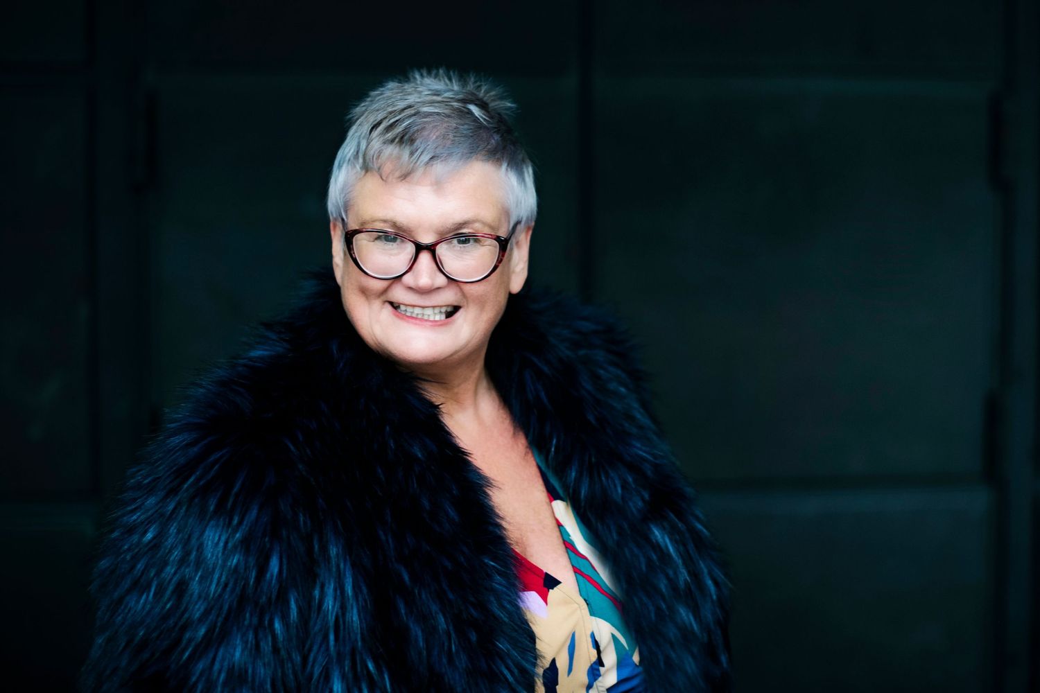 Smiling woman with short gray hair wearing a black fur coat photographed against a dark background.