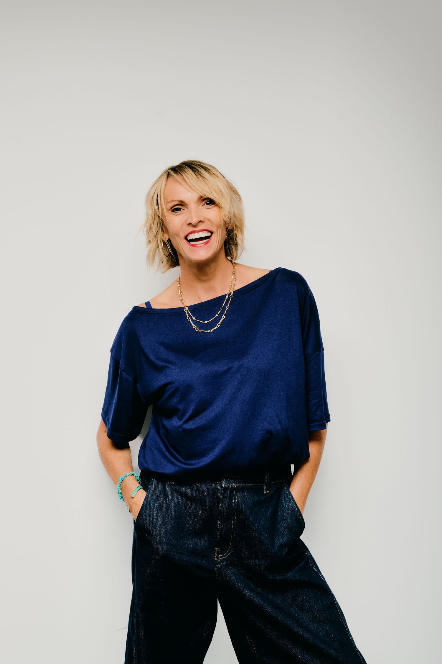 Smiling blonde woman in a navy blue top and dark pants posing confidently against a white background.