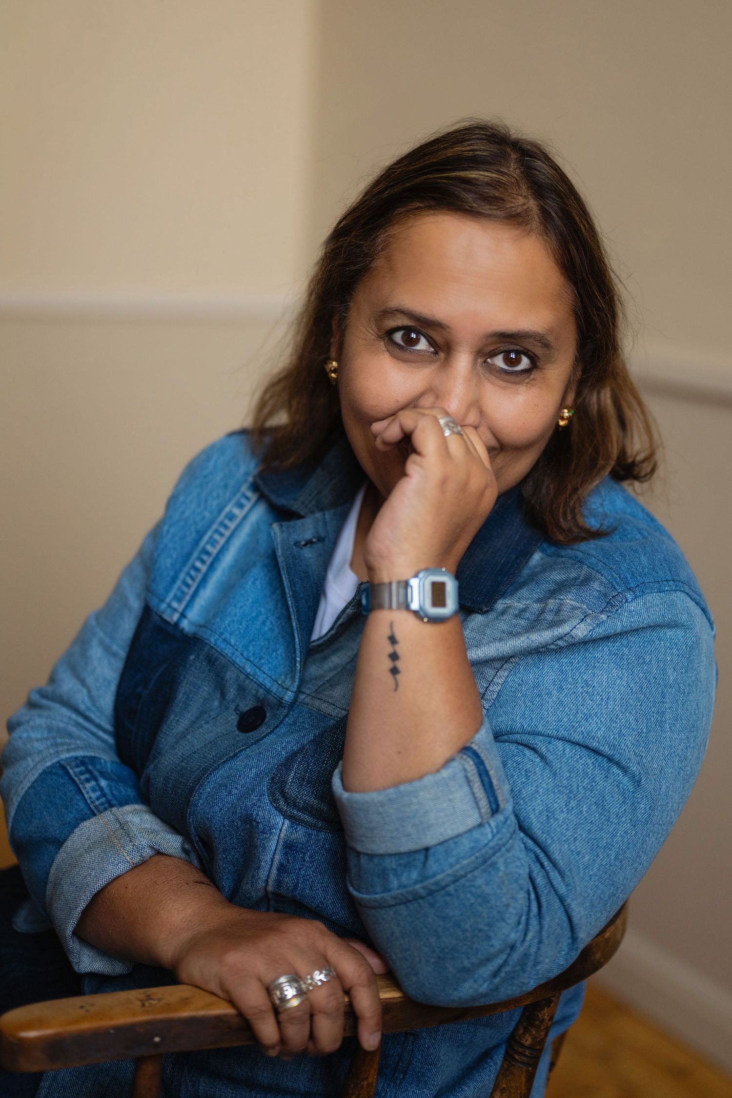 Woman in denim jacket sitting on a wooden chair with hand near face showing wrist tattoo.