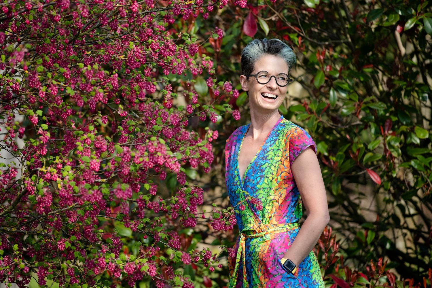 Woman with short teal hair in a colorful rainbow wrap dress standing beside pink flowering bushes outdoors.