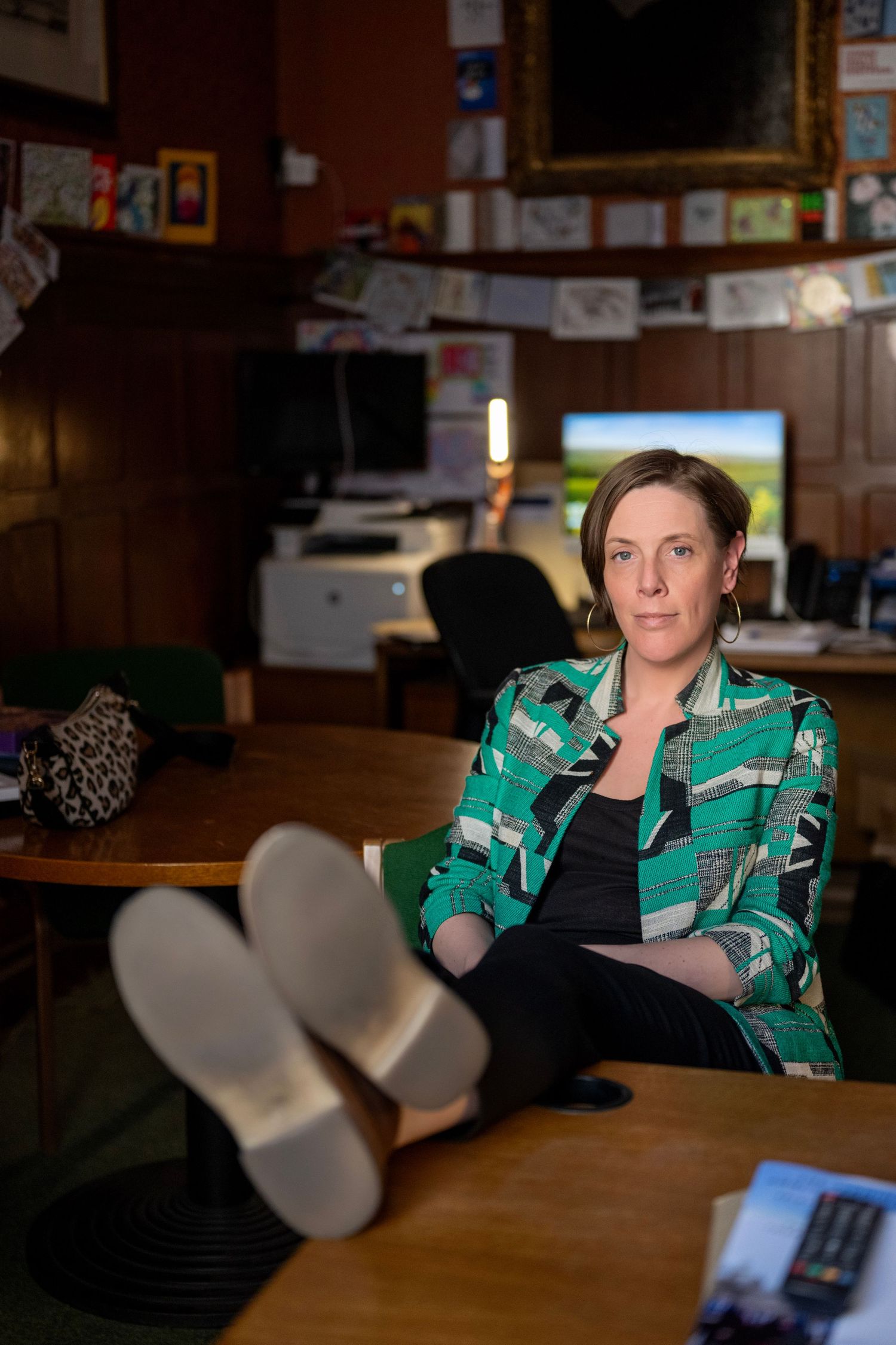 Woman in green patterned jacket sitting with feet on desk in cozy wood-paneled home office.