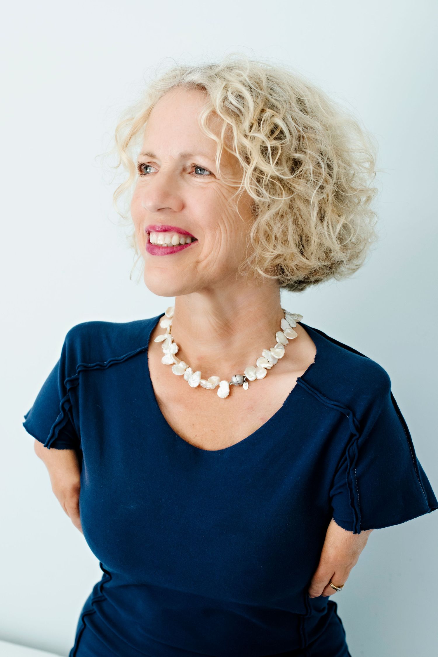 A smiling curly blonde-haired woman in a navy top and pearl necklace looks sideways against a light background.