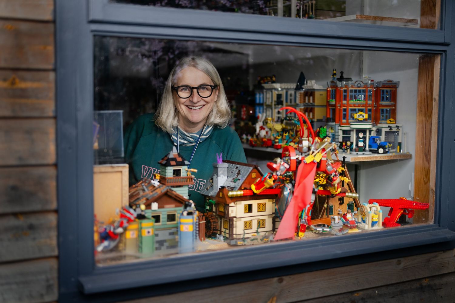 Smiling woman with blonde bob and glasses poses behind window with vibrant LEGO display collection.