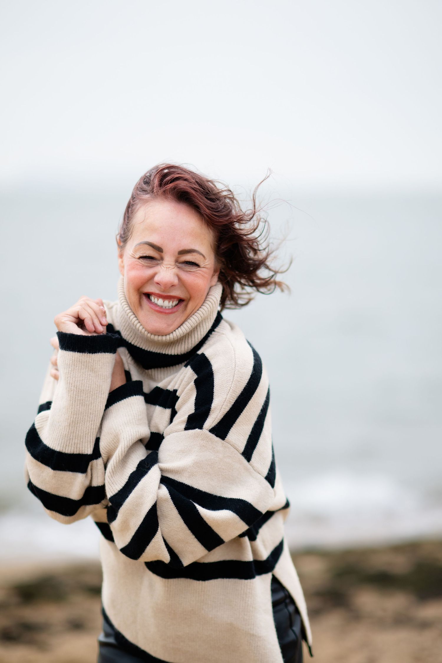 A joyful woman in a striped turtleneck sweater laughs on a windy beach.