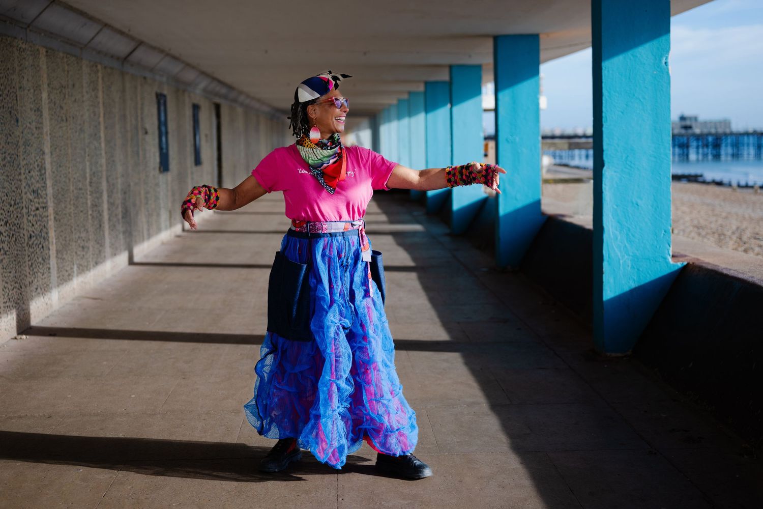 A woman in a pink top and blue ruffled skirt dances joyfully in a sunlit seaside colonnade.