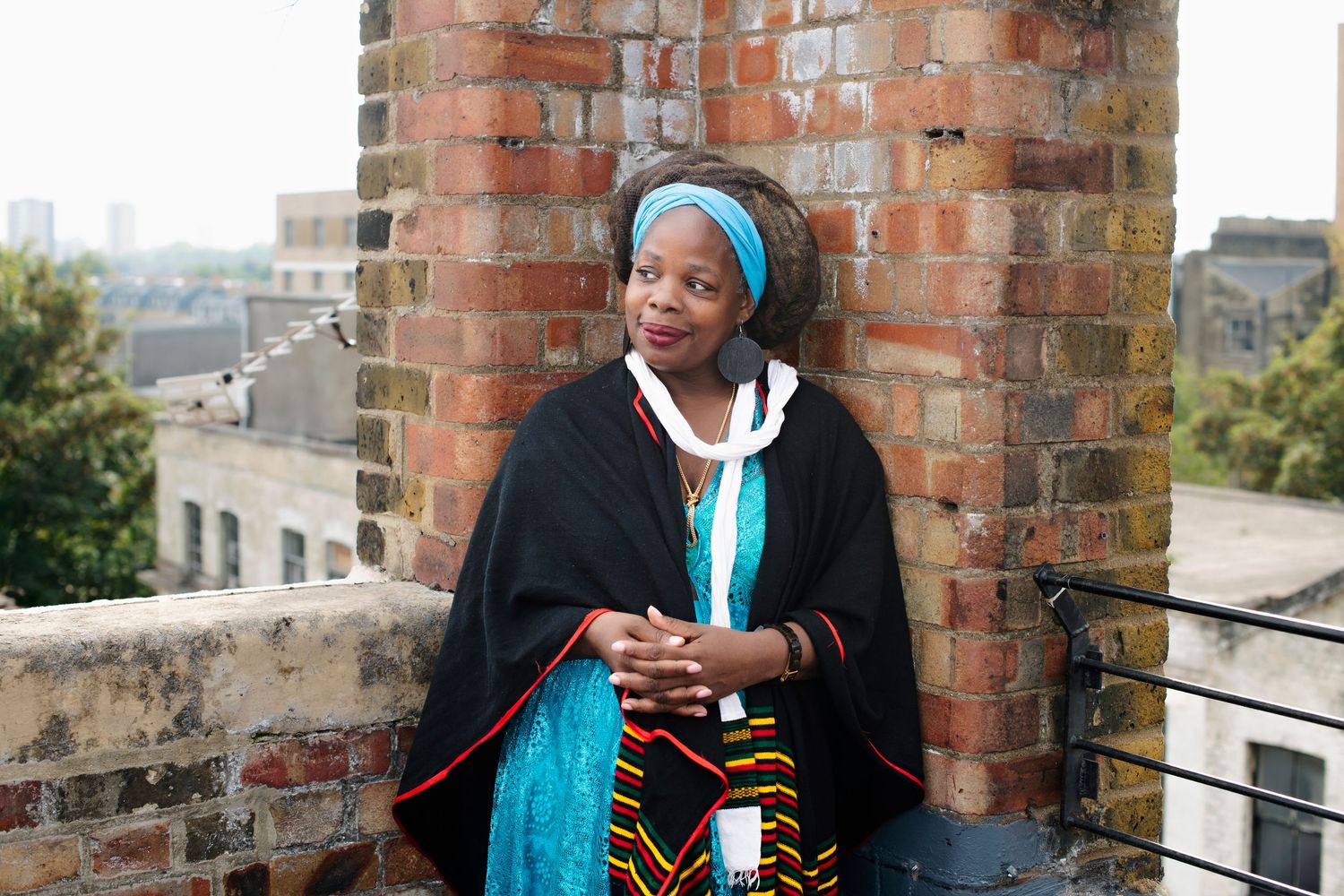 Woman in traditional African black cape with kente cloth stands on a rooftop terrace beside brick columns.