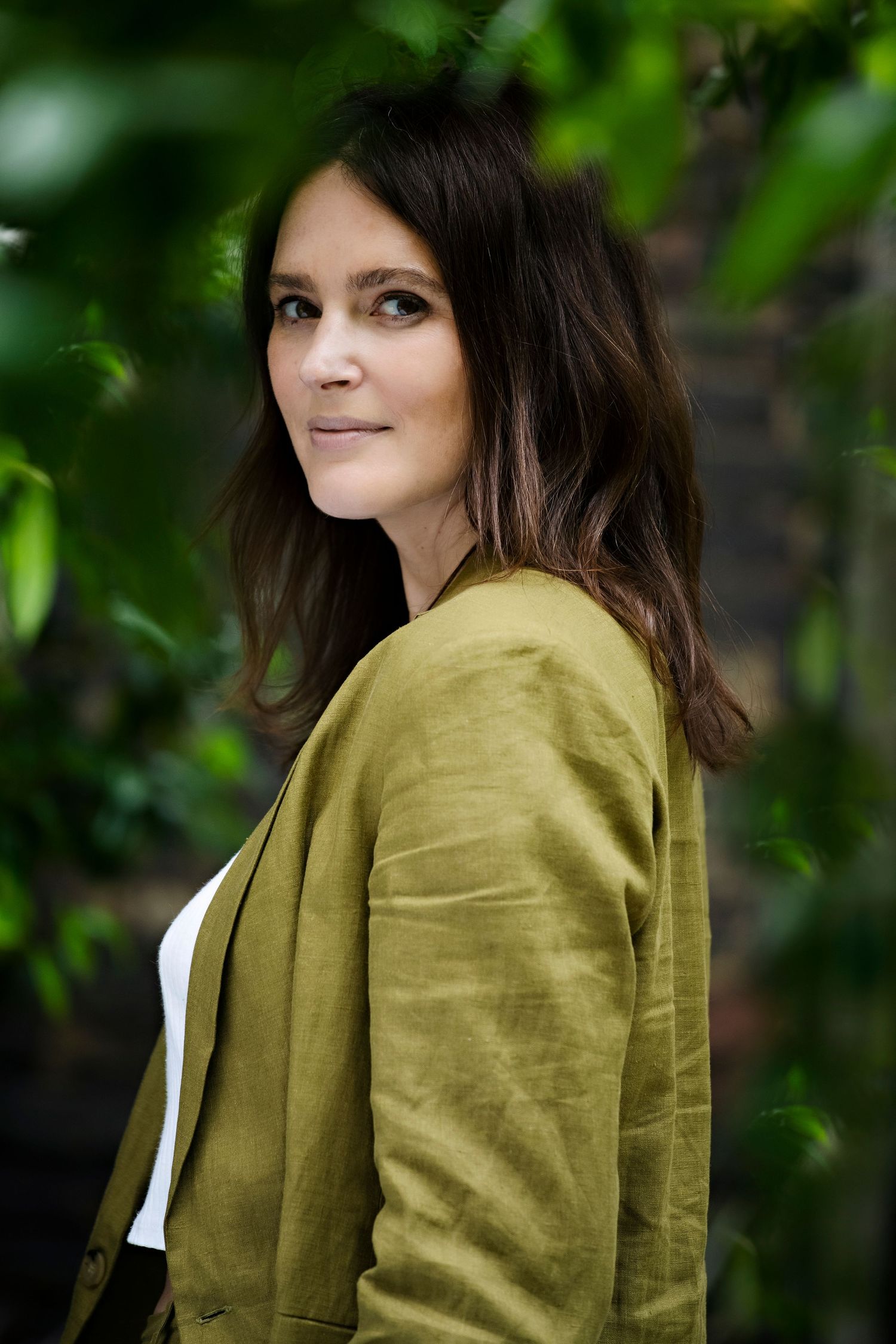 Brunette woman in an olive green jacket standing among lush green leaves in a garden setting.