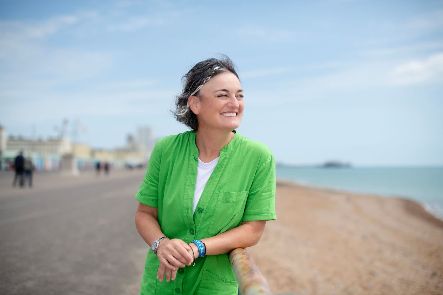 Smiling woman in green jacket enjoying a sunny day on a seaside promenade.