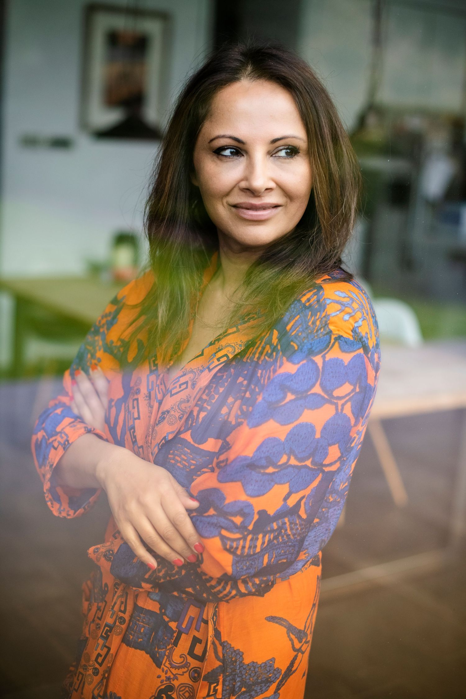 A confident woman in a colorful orange and blue patterned dress stands with arms crossed, smiling indoors.