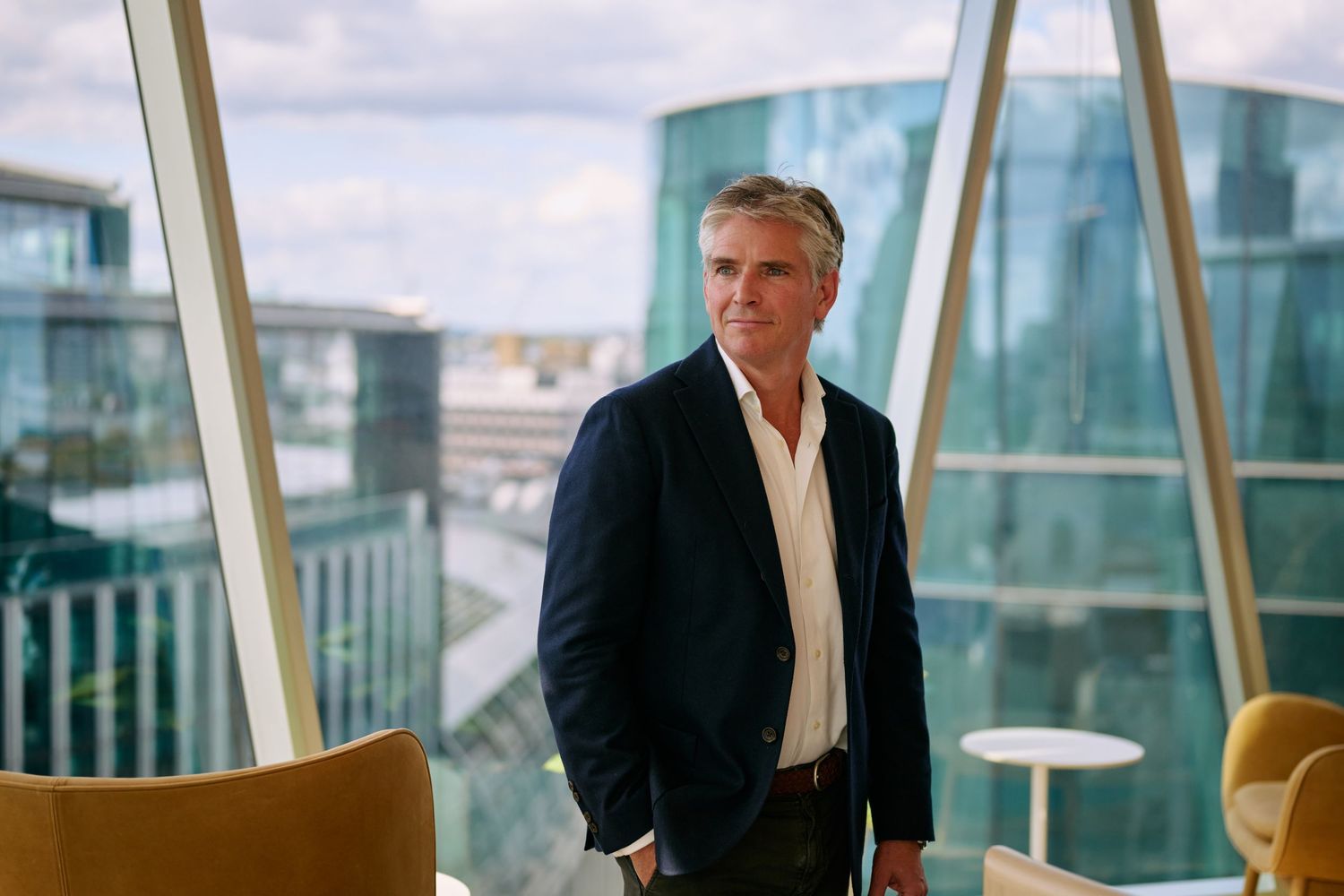 Professional businessman in navy blazer standing in modern glass office building with city views.