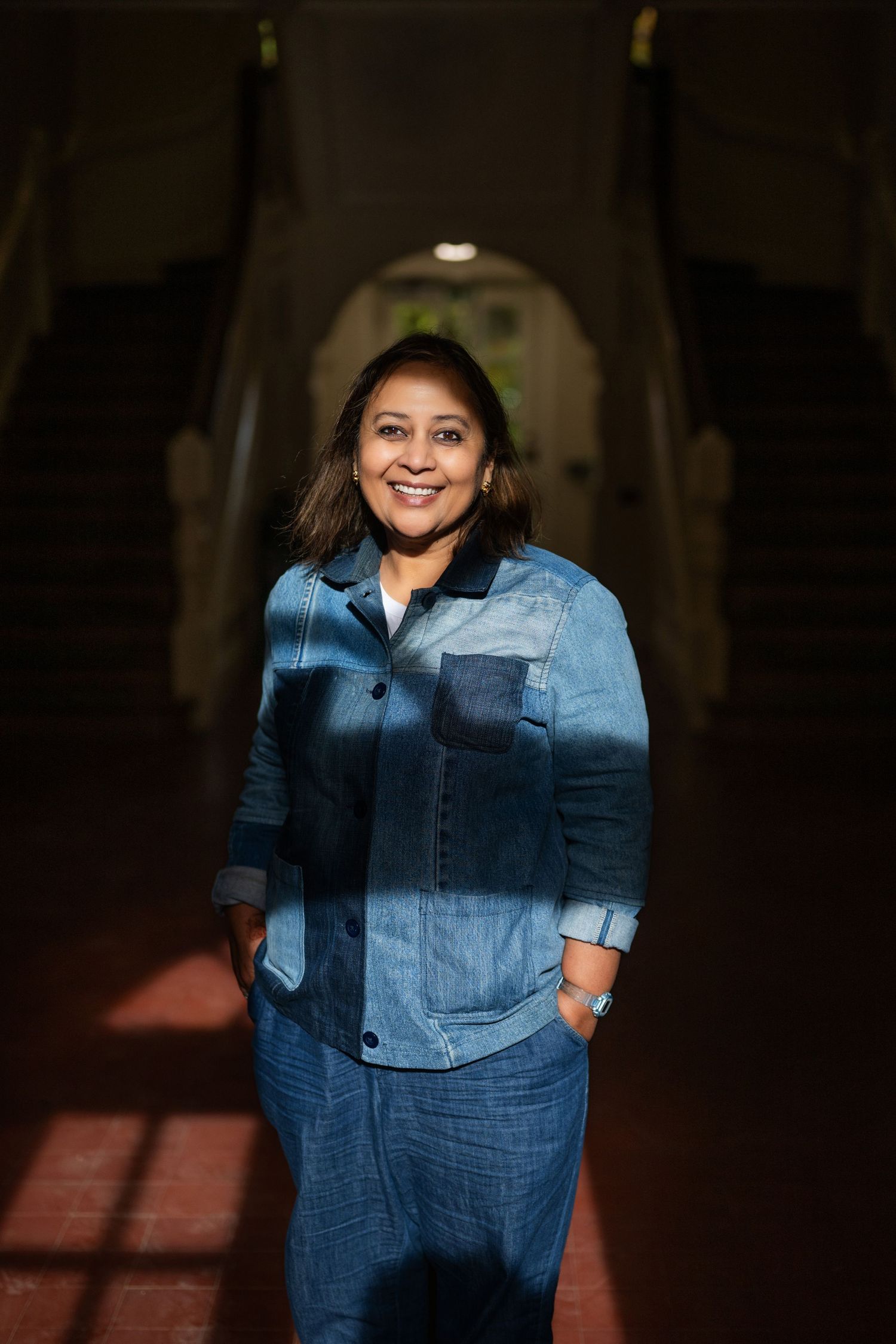 Woman in patchwork denim jacket and pants smiling in a dramatic arched hallway with warm lighting.