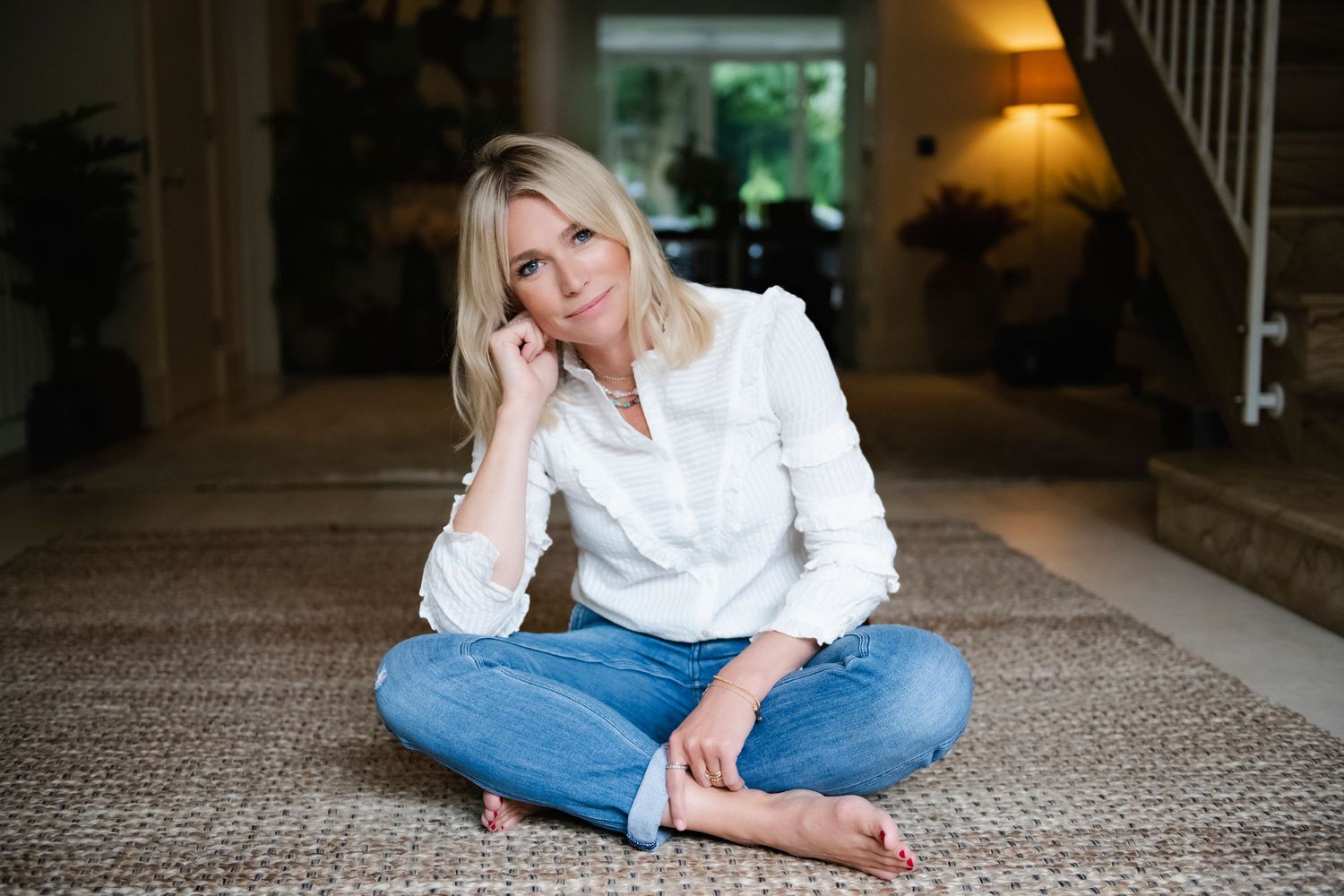 Blonde woman in white shirt and jeans sitting cross-legged on floor in elegant home interior.