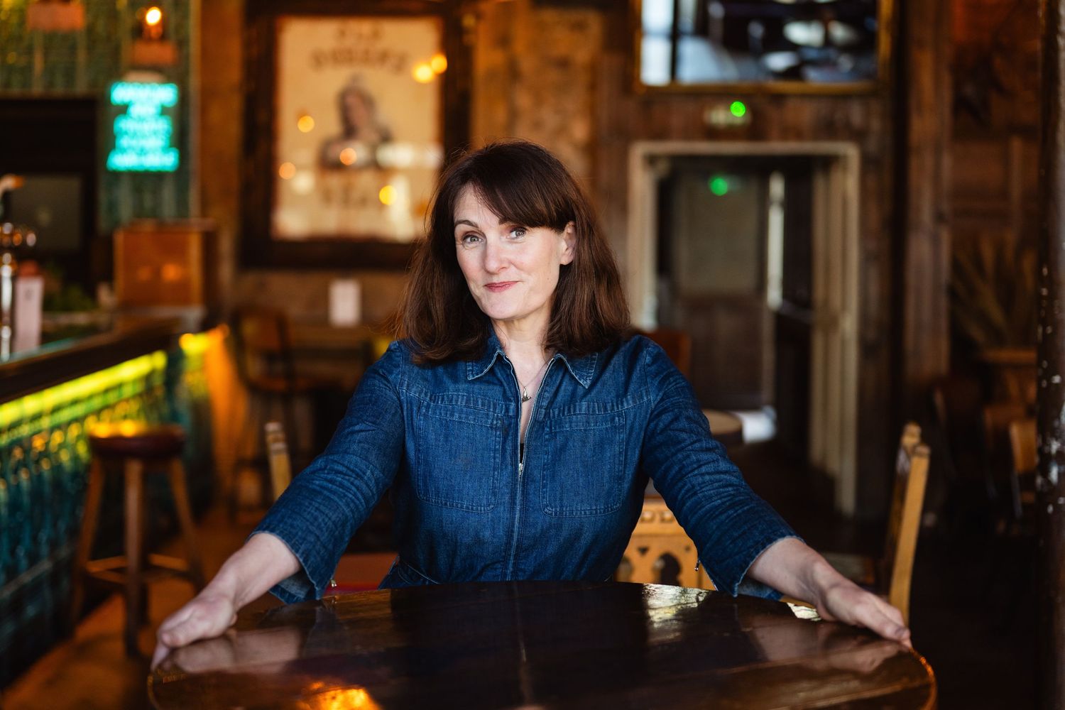 Woman in denim shirt seated at wooden table in rustic bar interior with warm lighting.