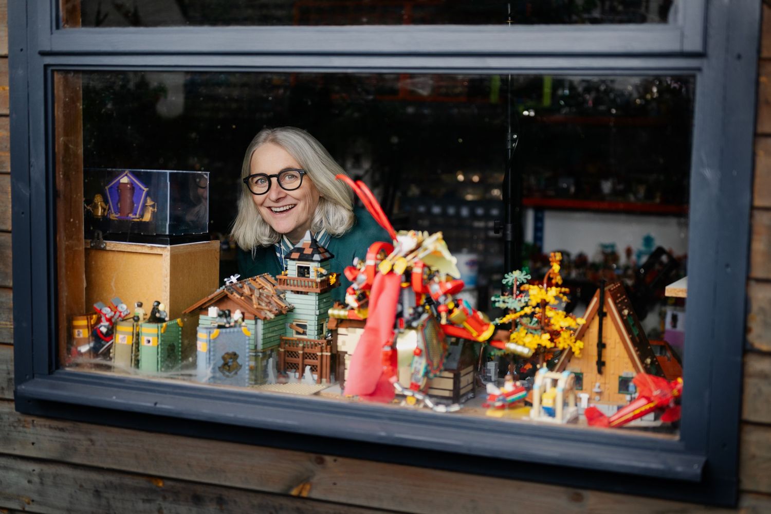 Woman with glasses peers through a window display filled with colorful LEGO sets and figurines.