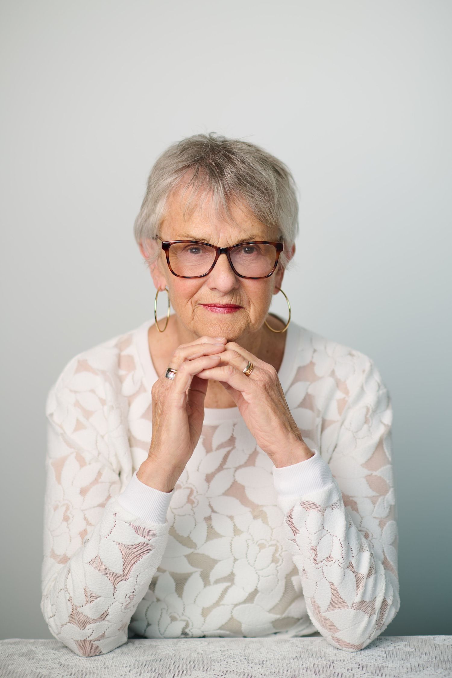 Elegant senior woman with short gray hair and glasses, wearing a white lace top, resting her hands under her chin.