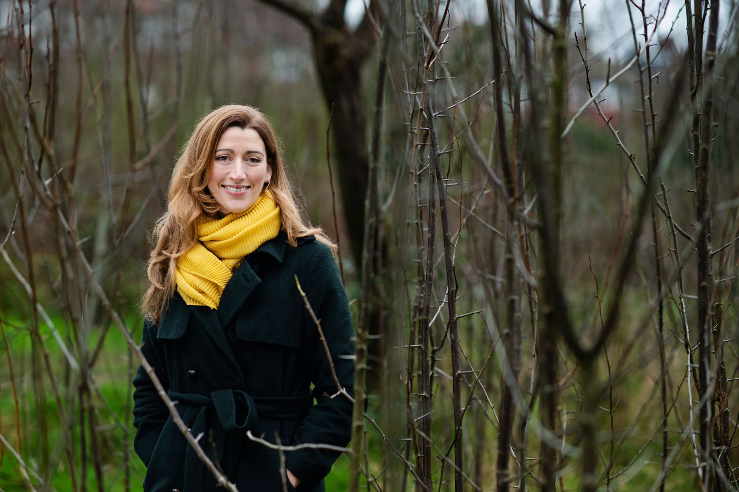 Smiling woman with long hair wearing yellow scarf and black coat standing among bare winter trees.