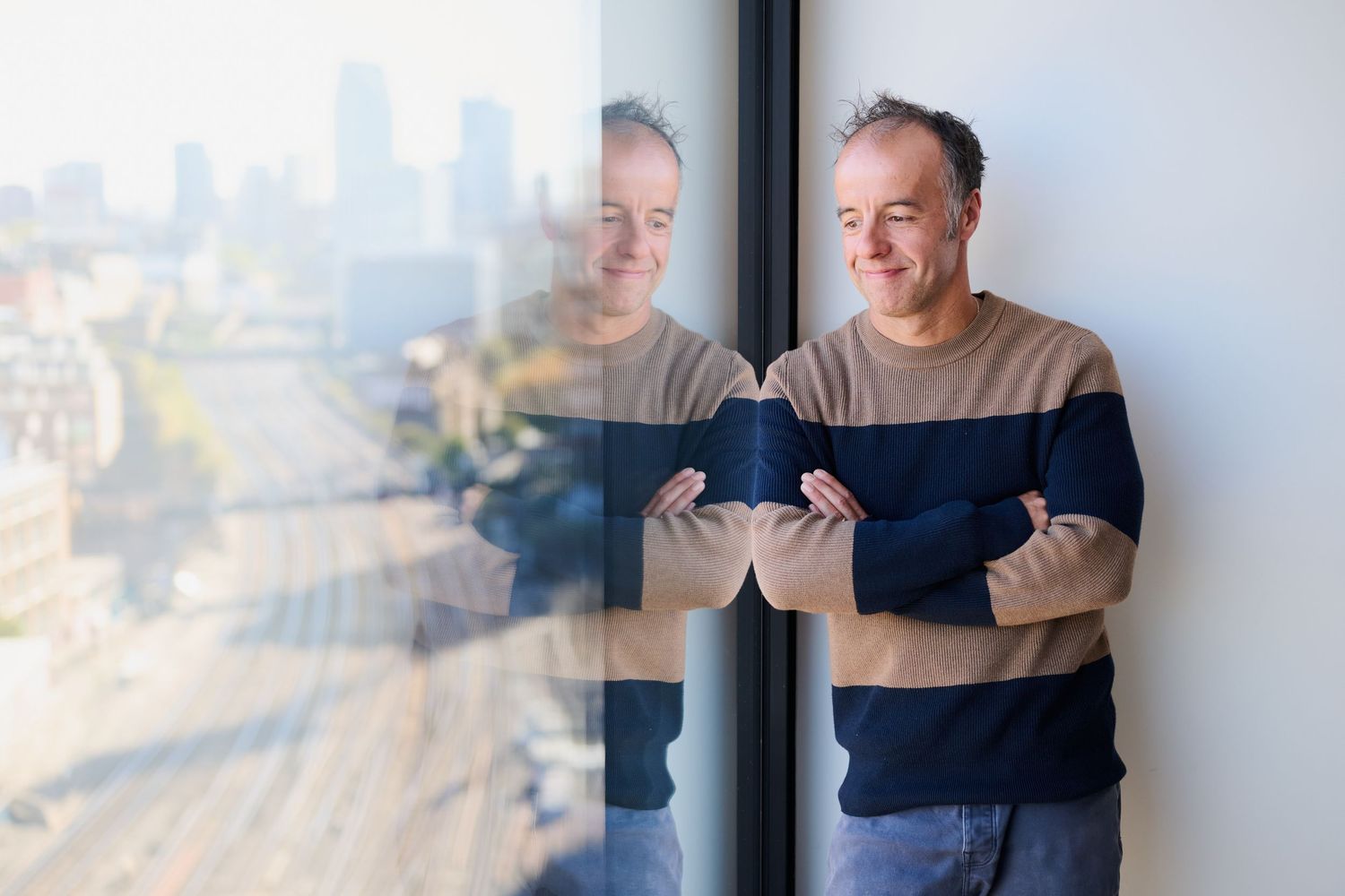 Relaxed middle-aged man in striped sweater leaning against window with city skyline view behind him.