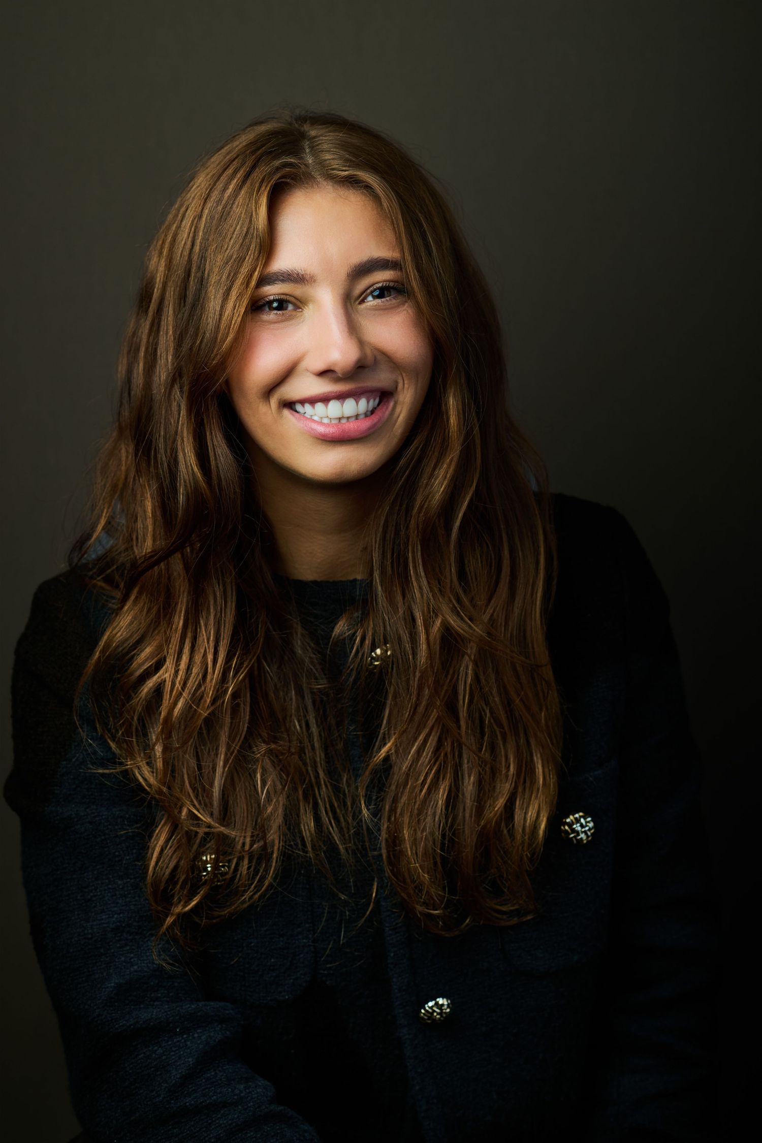 Young woman with long brown hair smiling confidently in a black blazer against dark background.
