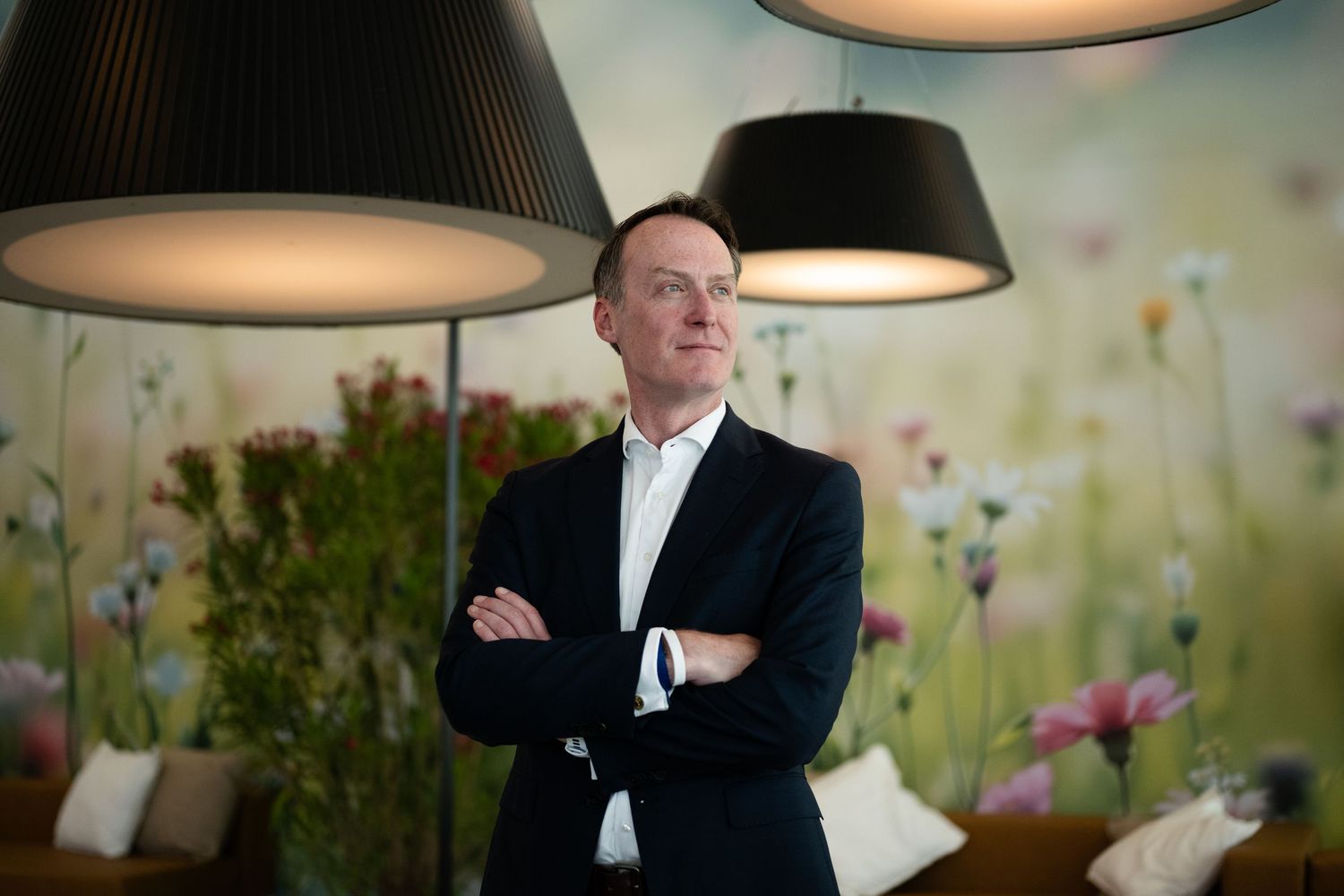 Man in black blazer with arms crossed standing beneath pendant lights with floral mural background.