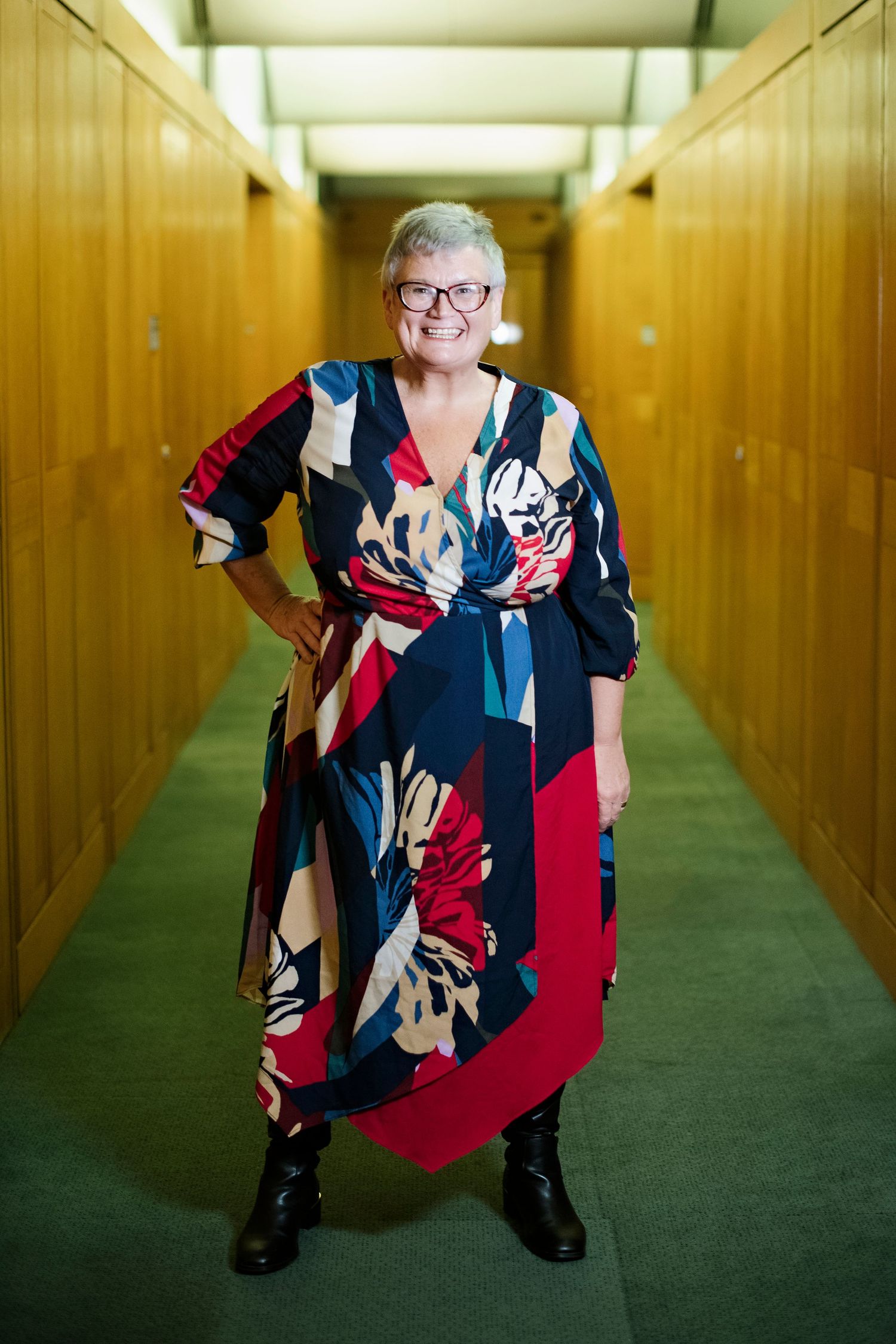 Smiling older woman with short gray hair and glasses in a colorful floral wrap dress posing in a hallway.