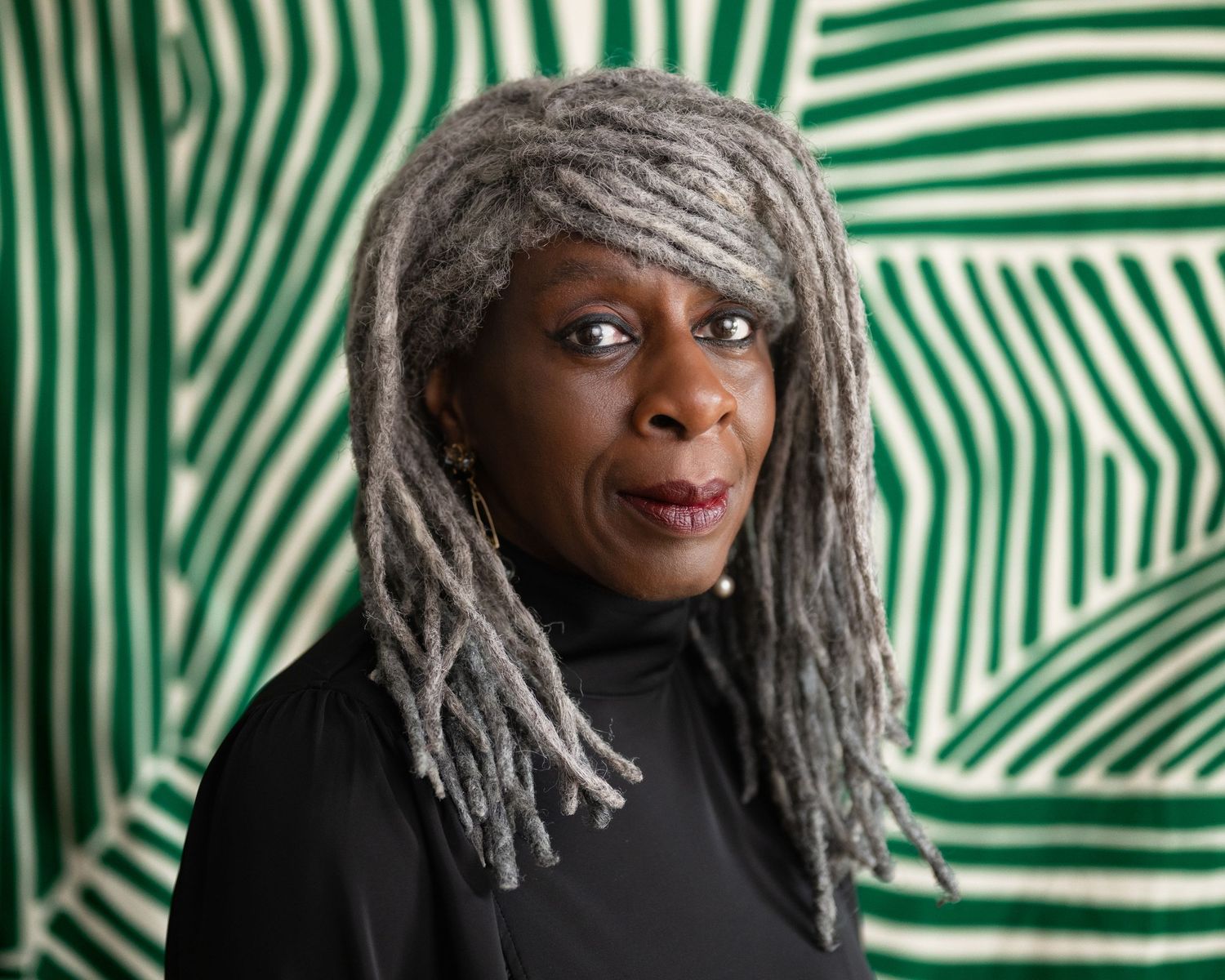 Woman with long gray dreadlocks wearing black turtleneck against green patterned background.