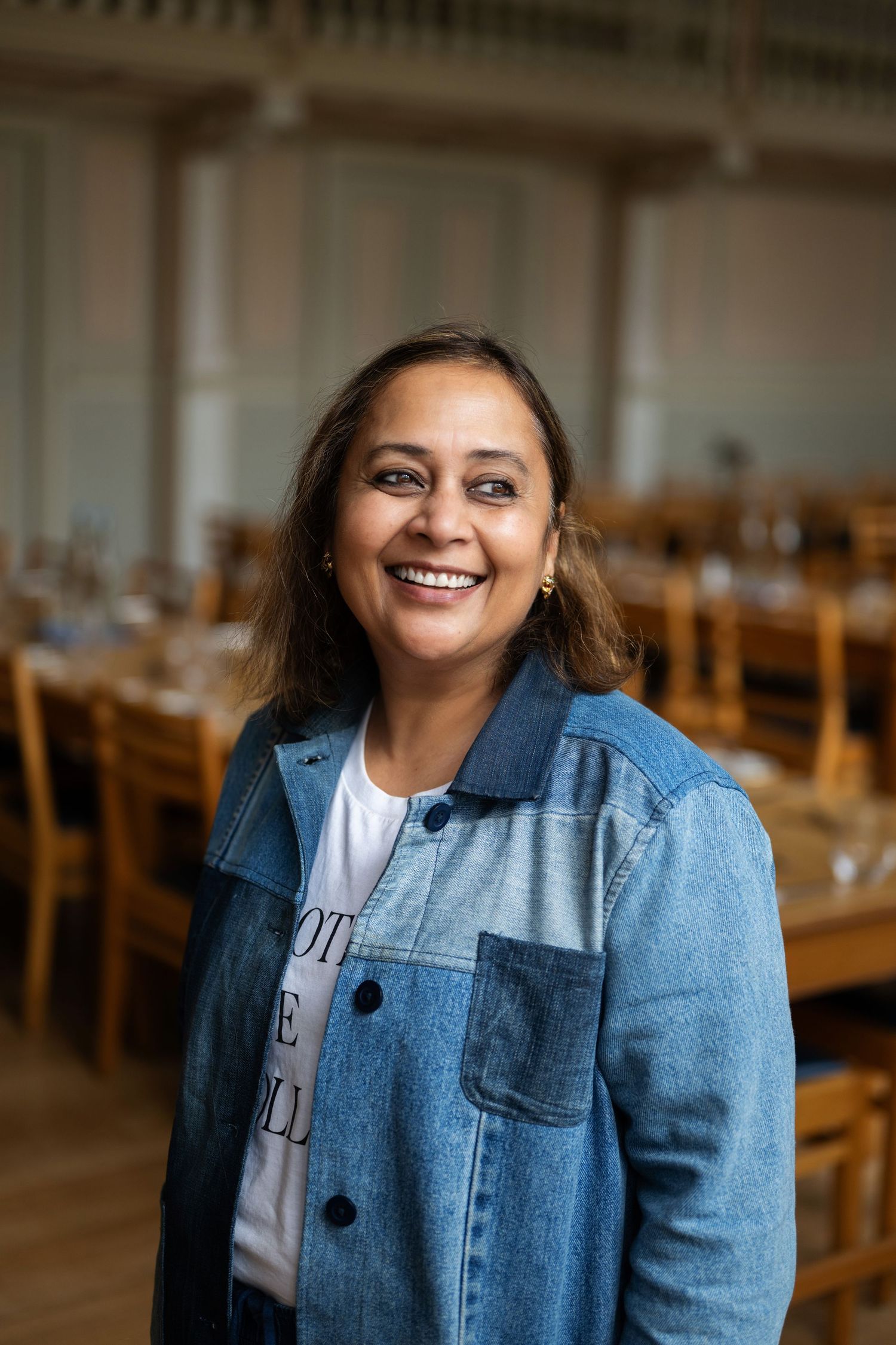 Smiling woman in patchwork denim jacket and white tee posing in a bright dining hall.