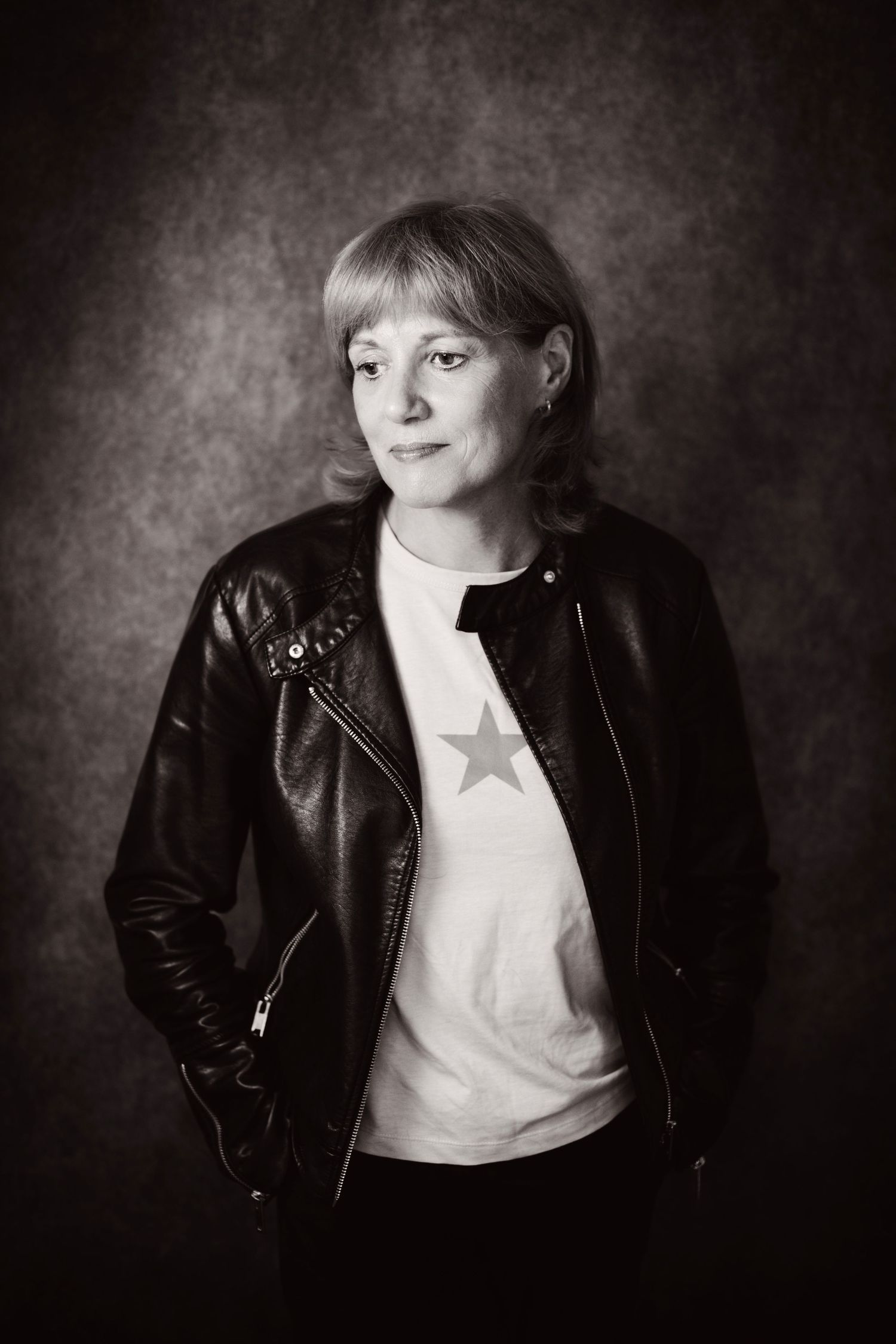 Black and white portrait of woman in leather jacket and star t-shirt standing against textured backdrop.
