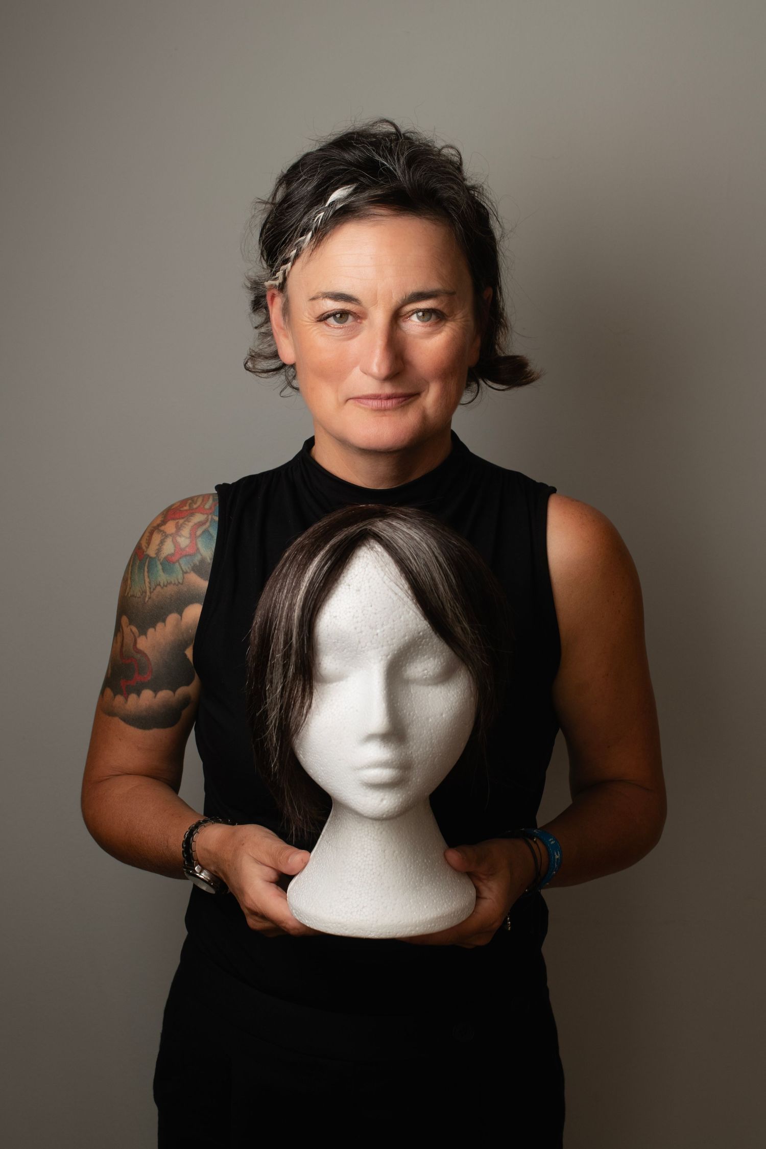Woman with tattoed arm holding a wig on a mannequin head against neutral gray background.