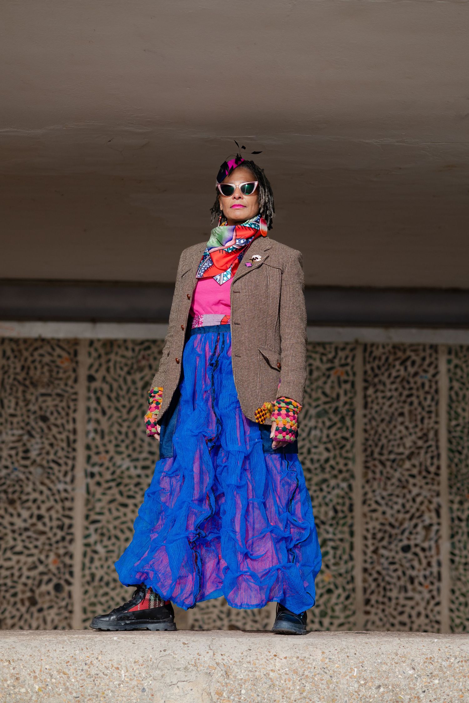 A boldly dressed woman in a blue skirt, tweed jacket, and colorful accessories stands under concrete overhang.