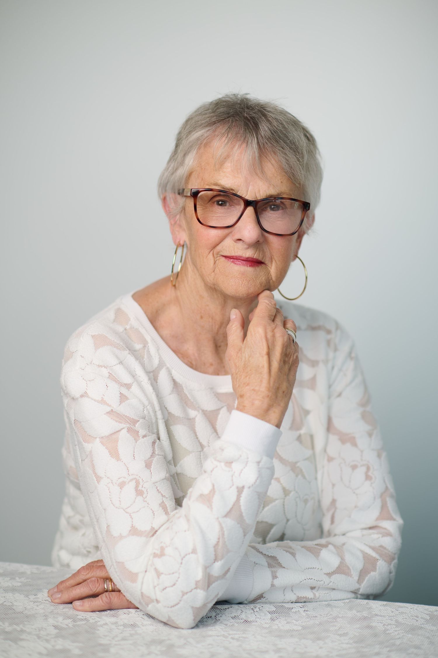 Elegant senior woman with short gray hair and glasses wearing a white lace top, posing thoughtfully at a table.