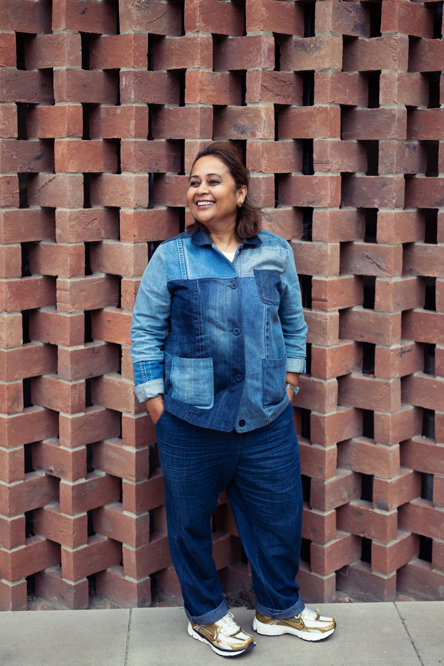 Smiling woman in patchwork denim outfit standing confidently against a decorative red brick wall.
