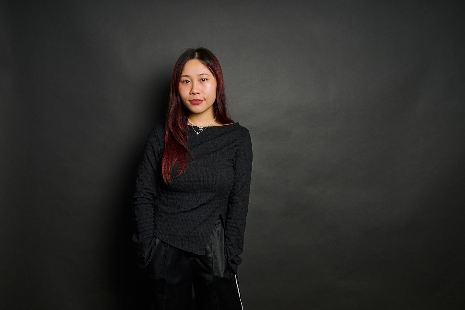Young Asian woman with red-highlighted hair wearing a black long-sleeve top, posing against a dark gray background.