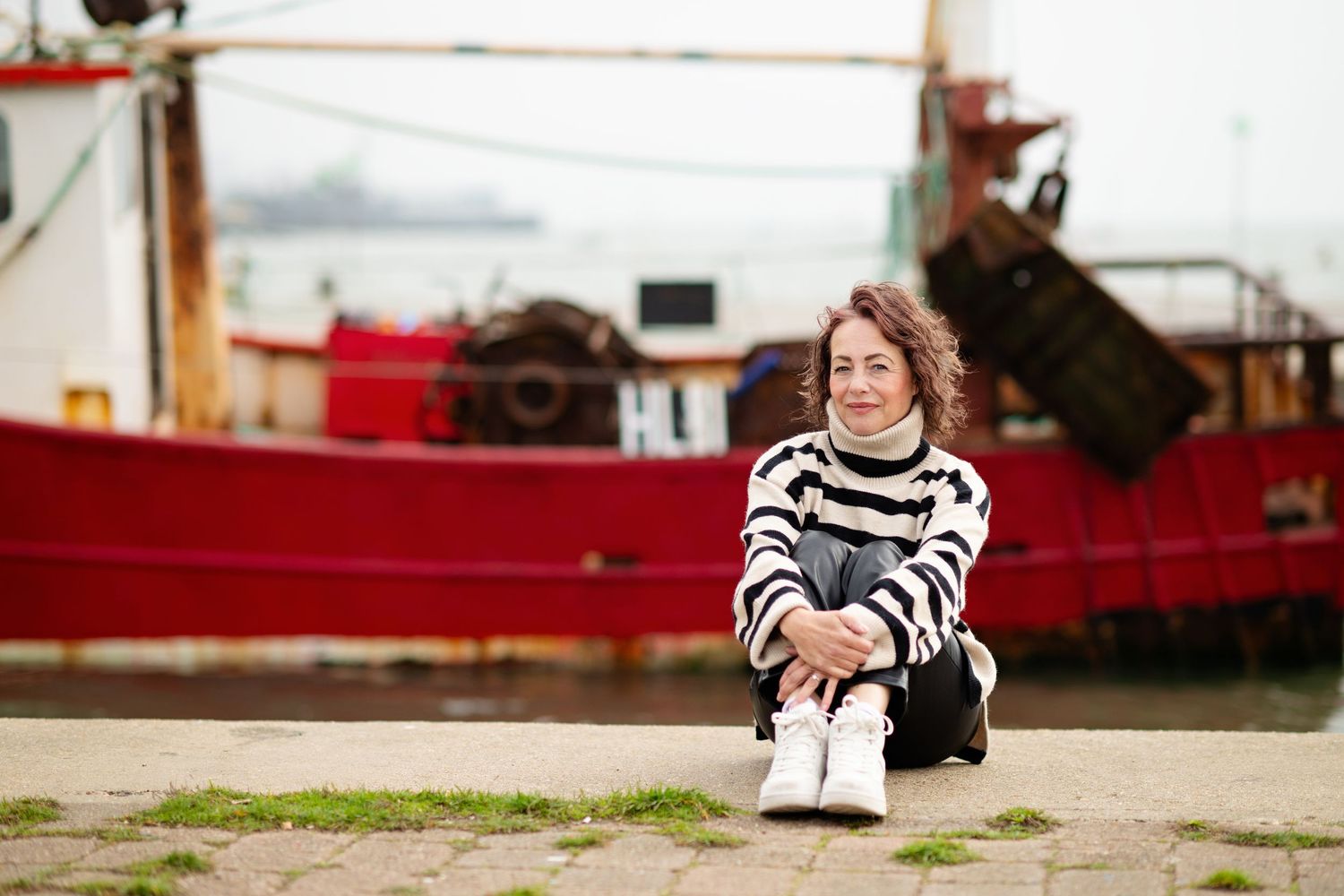 Woman in striped sweater sitting on harbor ground with red fishing boat in background.