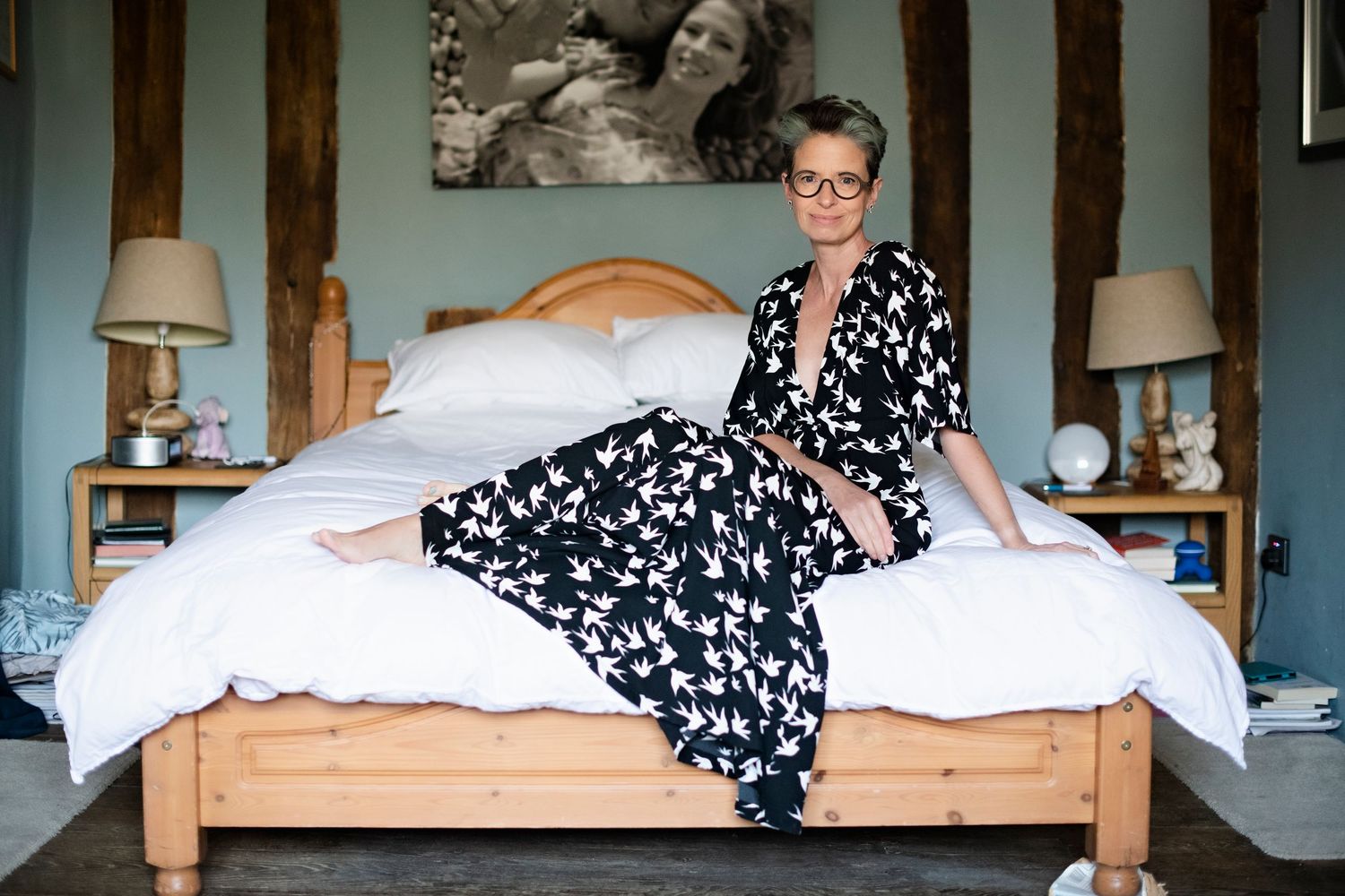 Woman in bird-print pajamas sitting on white bed with glasses in rustic bedroom setting.