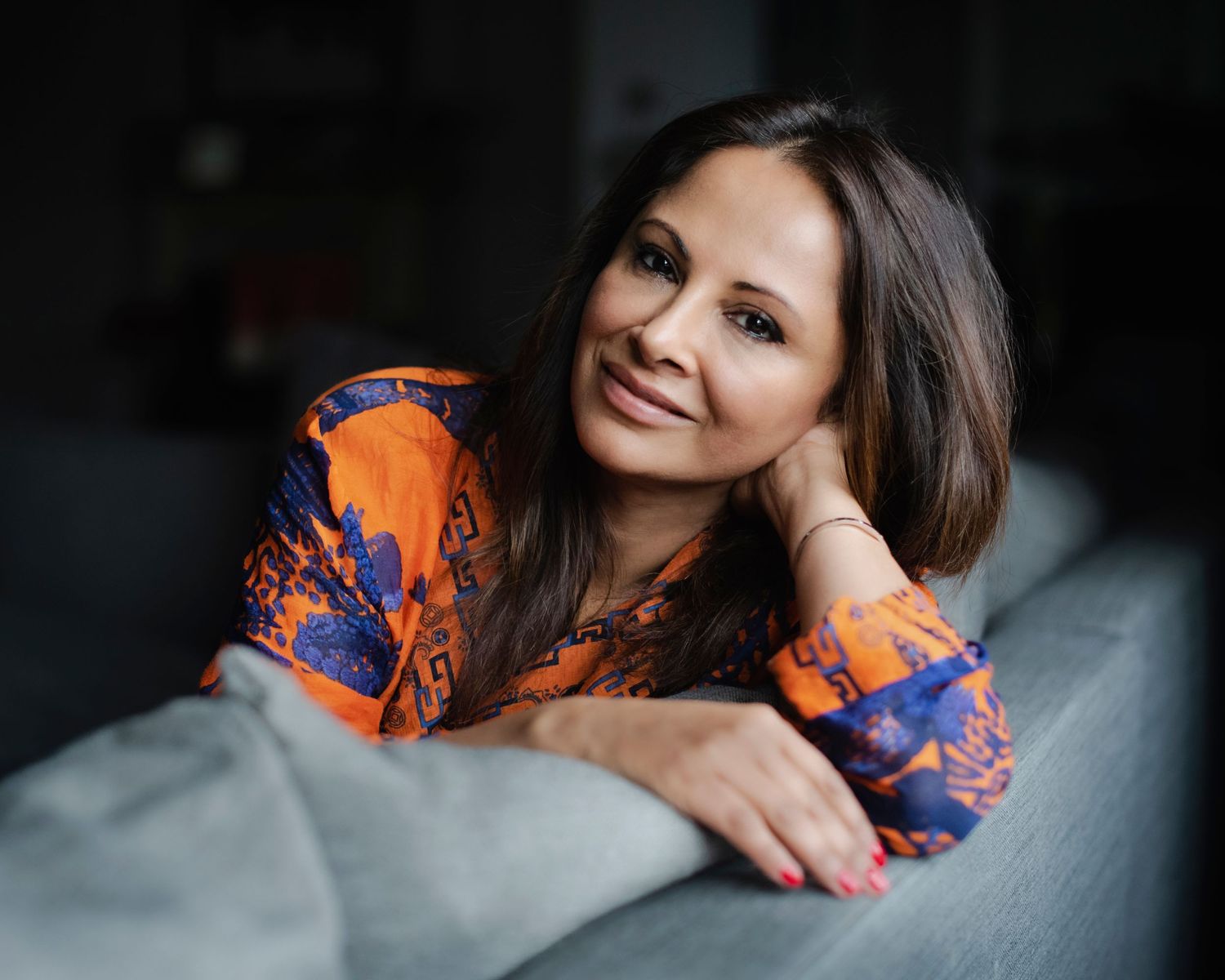 Smiling woman with brown hair in an orange floral blouse, relaxing on a gray sofa in a cozy indoor setting.