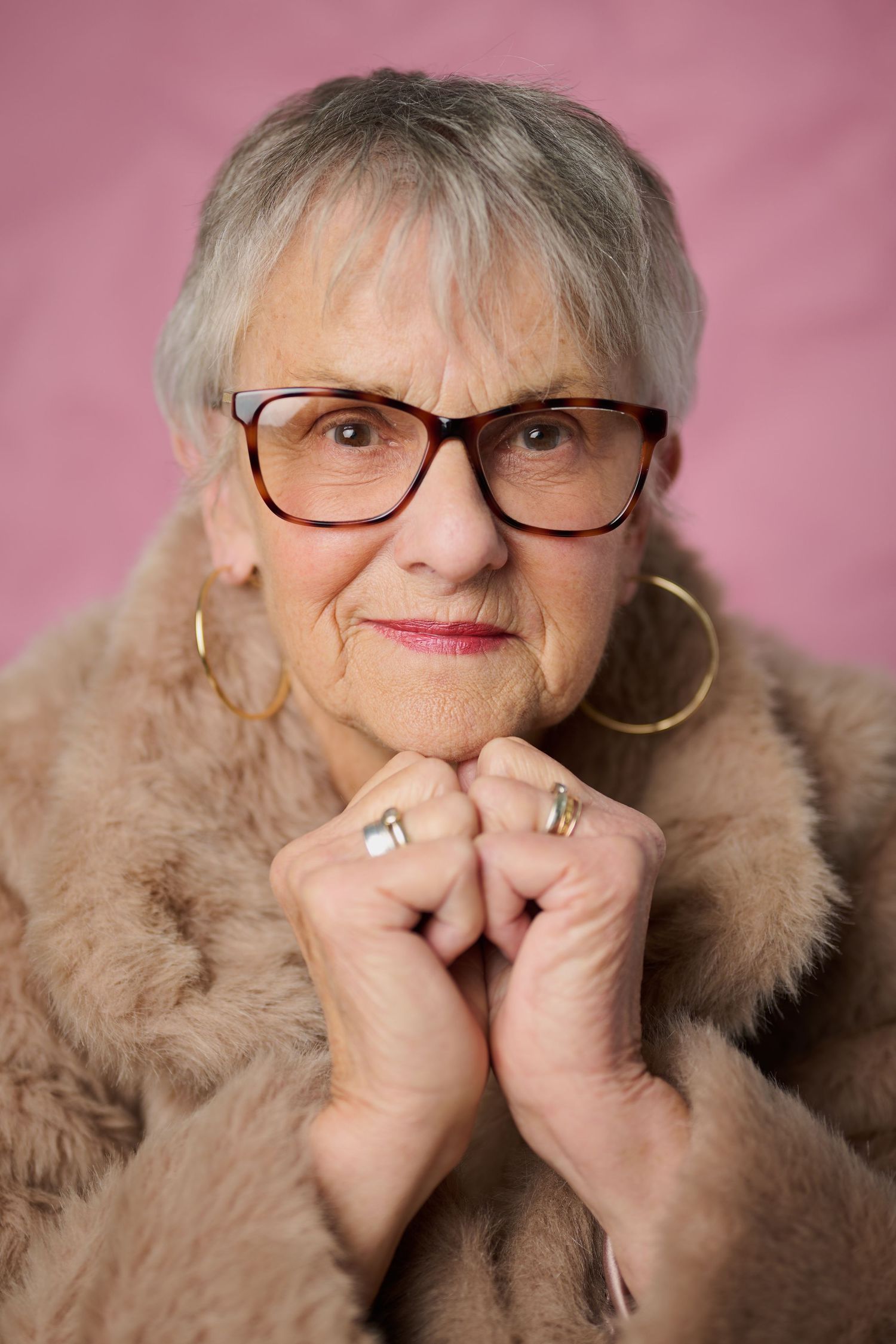 Elegant senior woman in fur coat and glasses with gold hoop earrings poses against pink background.