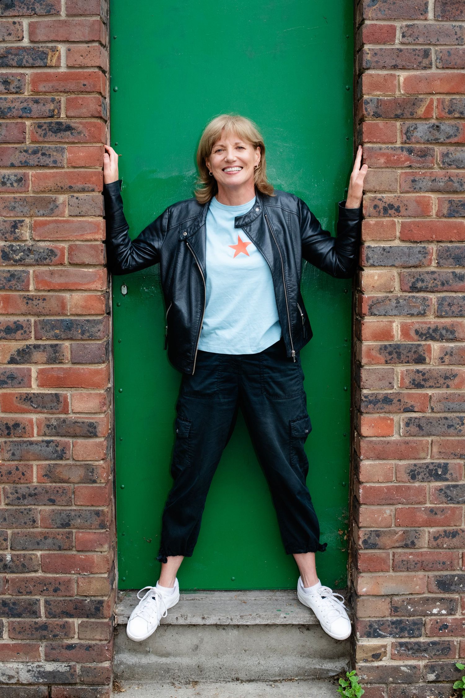 Cheerful woman in leather jacket and star t-shirt posing in green doorway framed by brick wall.