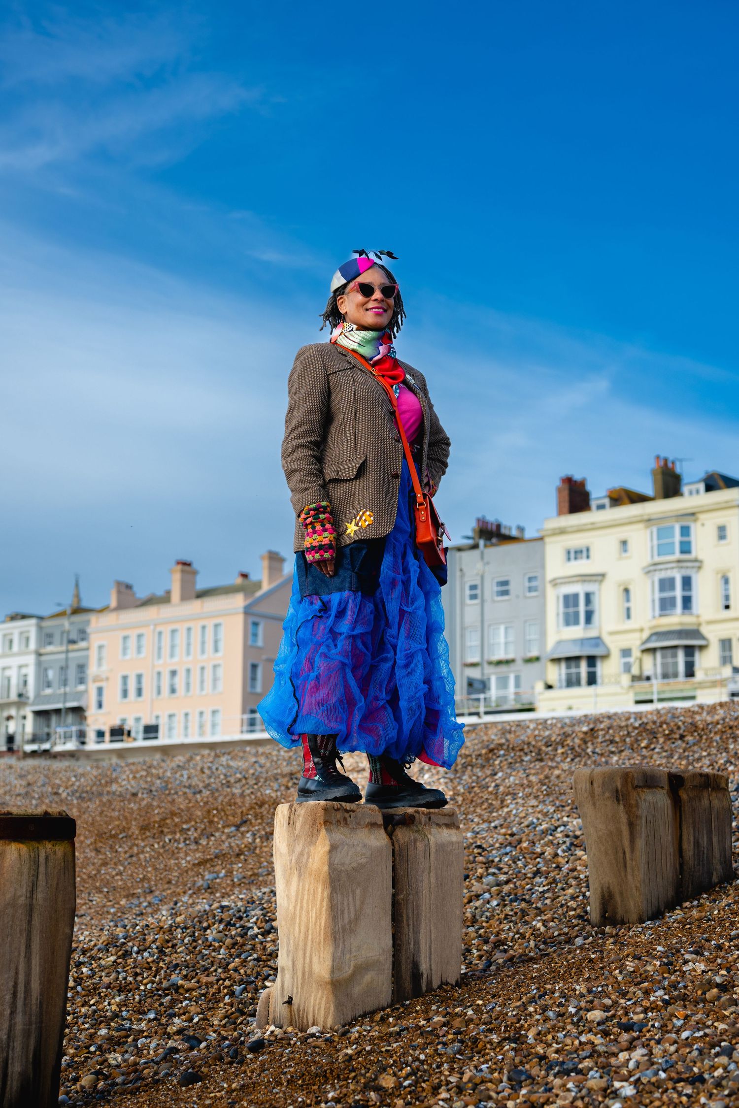 A woman in a vibrant blue skirt and brown jacket stands on wooden beach groynes under blue sky.