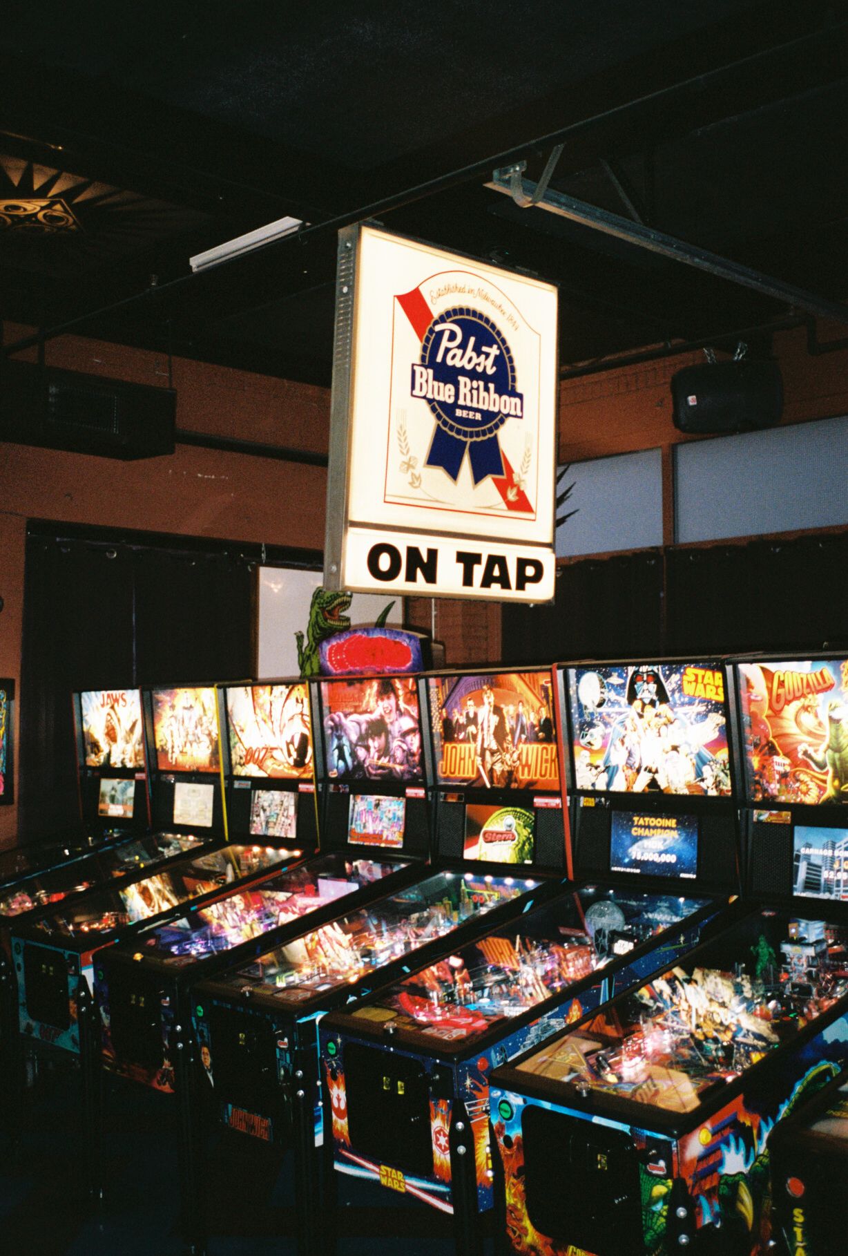 Retro arcade room with multiple pinball machines and a vintage Pabst Blue Ribbon 'On Tap' sign illuminated above.
