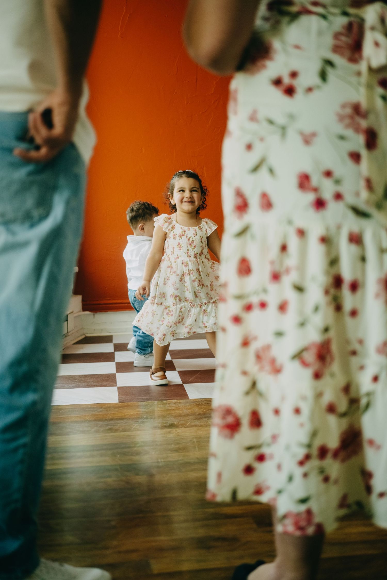 A smiling little girl in a floral dress stands on a checkered floor between adults in a room with an orange wall.